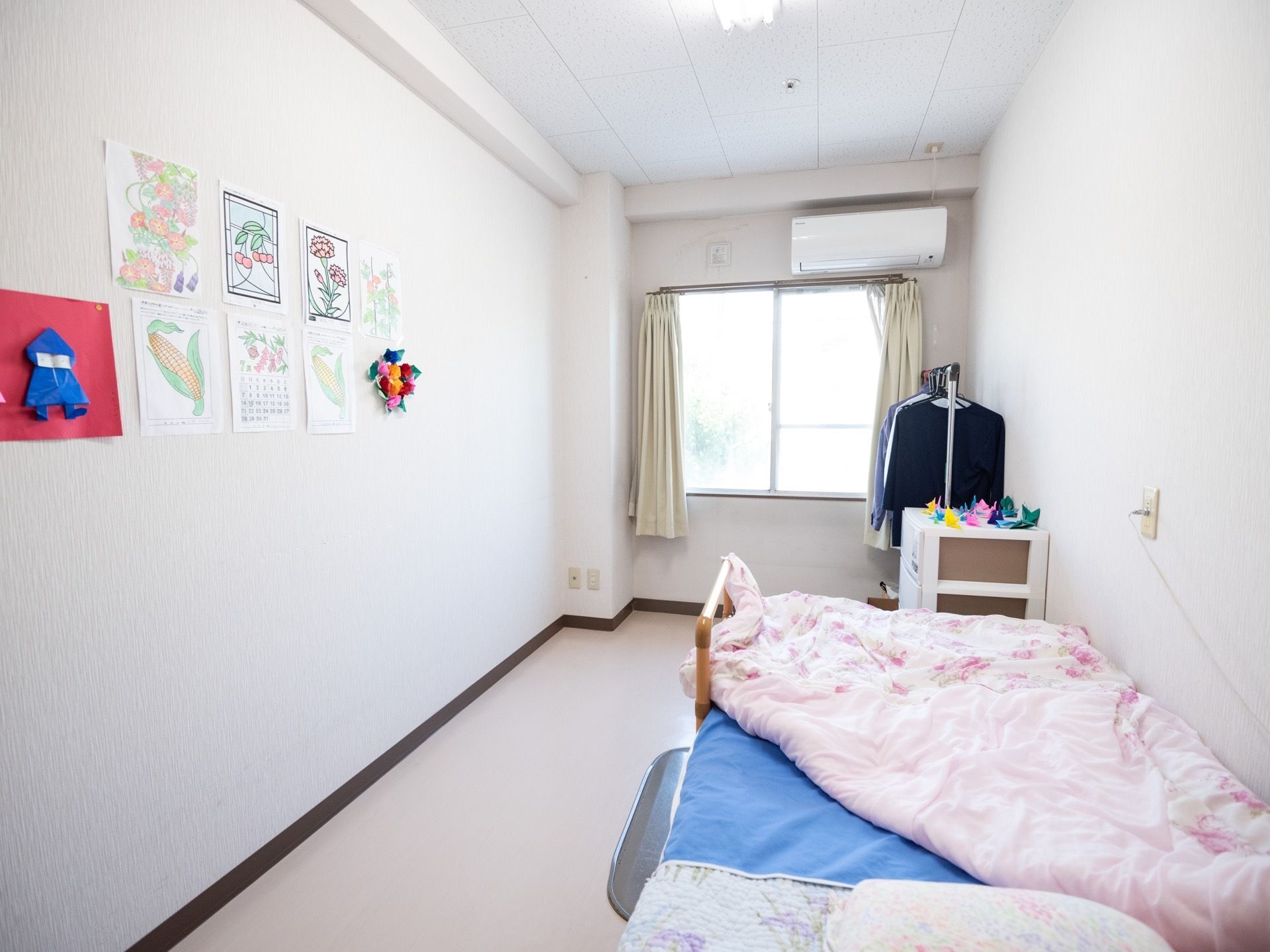 奥行きがあり、開放的な居室が完備されてある。窓から外の景色が見渡せる。ハンガーラックやベッドなどが置かれている。