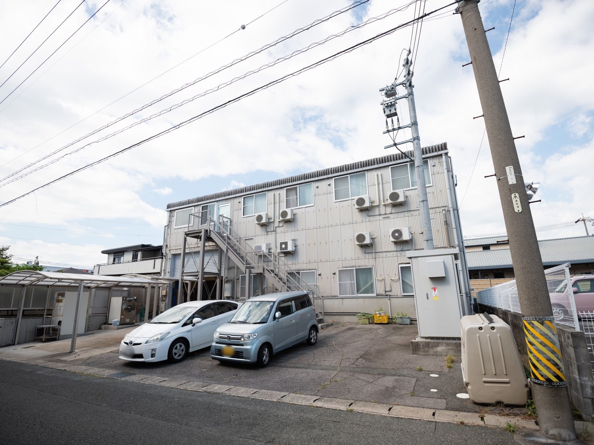 ワンカラーのシンプルな外観である。建物前に駐車場が完備され、車が２台停まってある。真正面は道路が走っている。