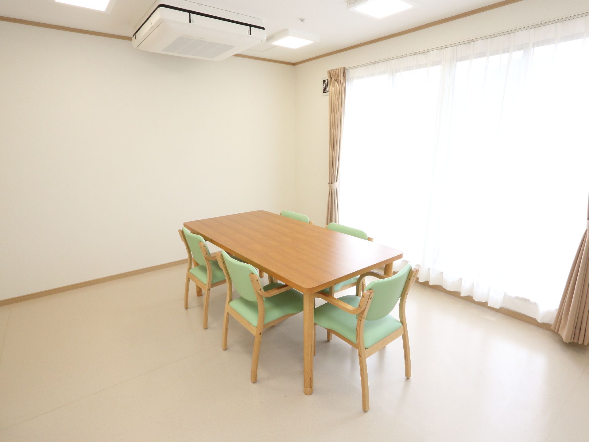 テーブルと椅子はウッド調である。椅子の座る部分や背もたれは緑色である。白とピンクのカーテンがある。天井には大きなエアコンが設置されている。