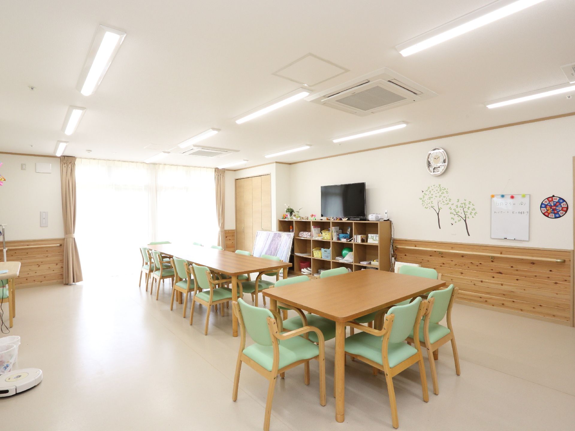 みんなで食事できるように、テーブルと椅子が設置されている。時計や大型テレビ、ウッド調の棚もある。明るく広々としている。