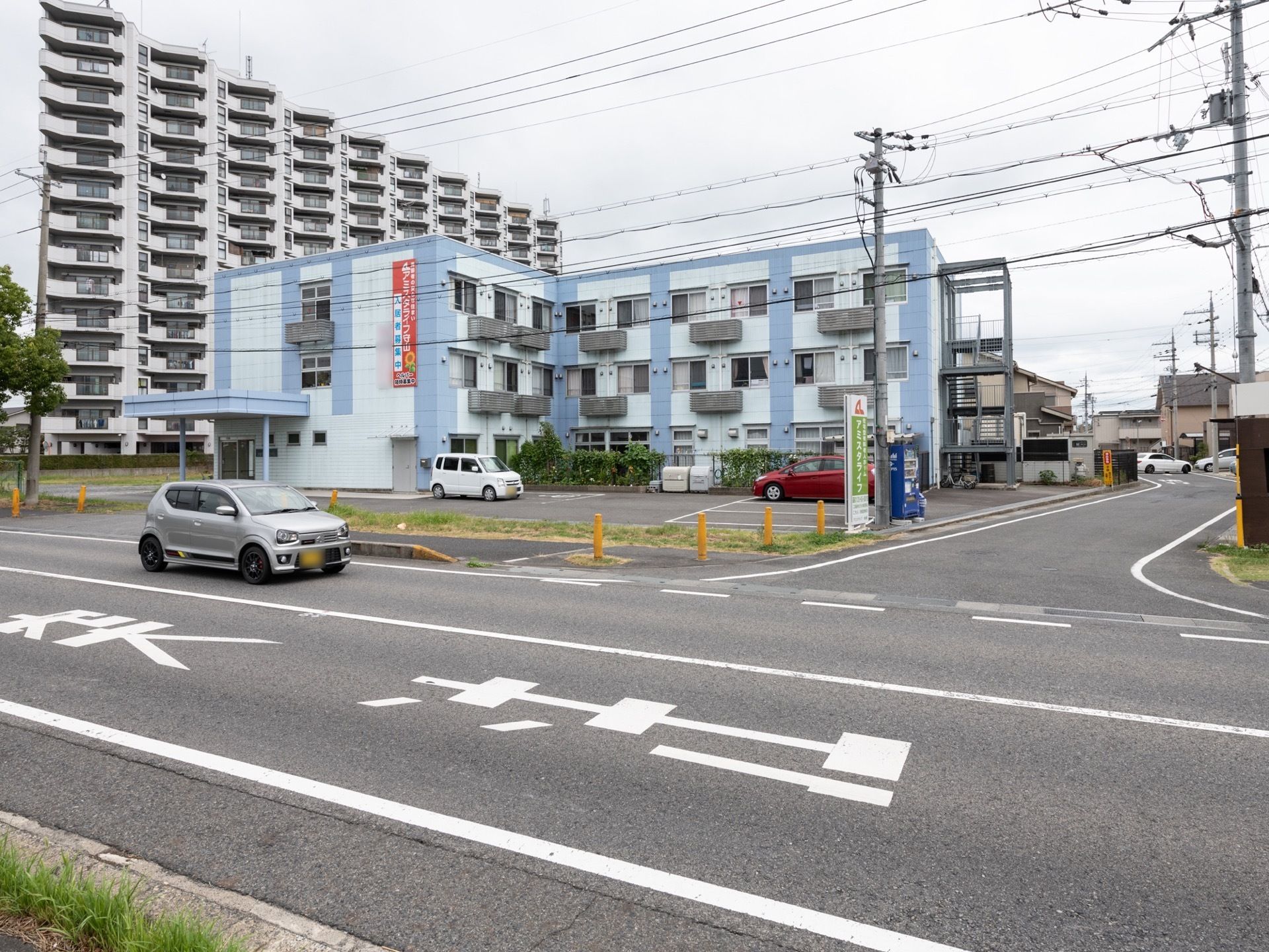 Ｌ字型の3階建てで、水色の外壁の施設である。隣には駐車スペースがあり、飲み物の自動販売機も設置されている。