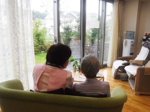 ソファーに寄り添って座り、ガラス越しに庭を見ている高齢の女性とスタッフ