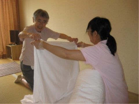 スタッフに手伝ってもらいながら笑顔でシーツをたたむ高齢の女性