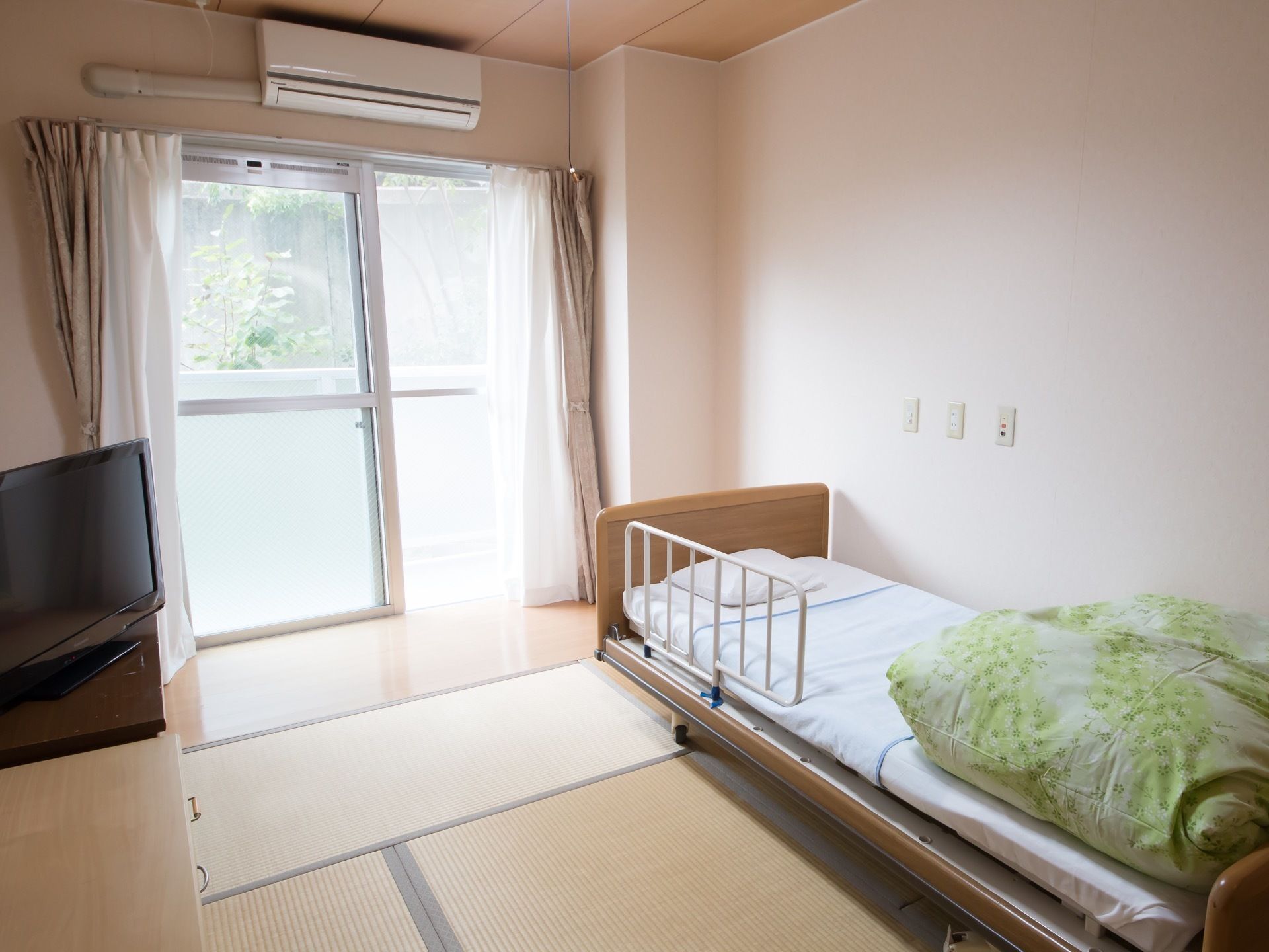 和室の居室である。低床の介護用ベッド、エアコン、テレビ、チェストが置いてある。窓は掃き出し窓で、明るい。