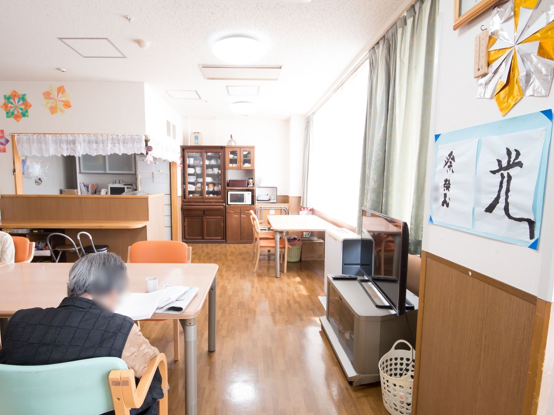 共有スペースにはテーブル、椅子、テレビが設置され、対面式のキッチンがあり、奥には食器棚が見える。壁には入居者様の作品が展示されている。