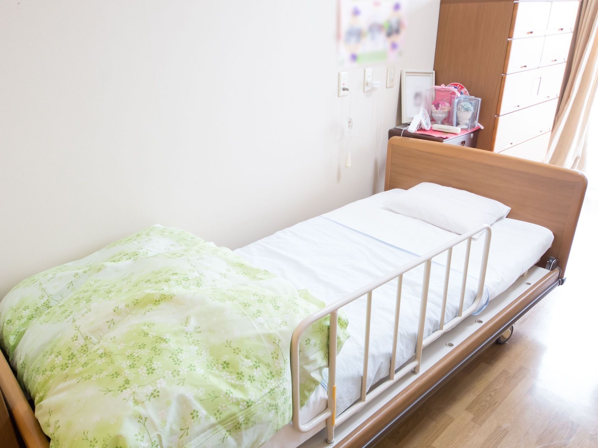 フローリング仕様の居室である。低床の介護ベッドが設置され、奥にはチェストがあり、置物や写真が飾ってある。