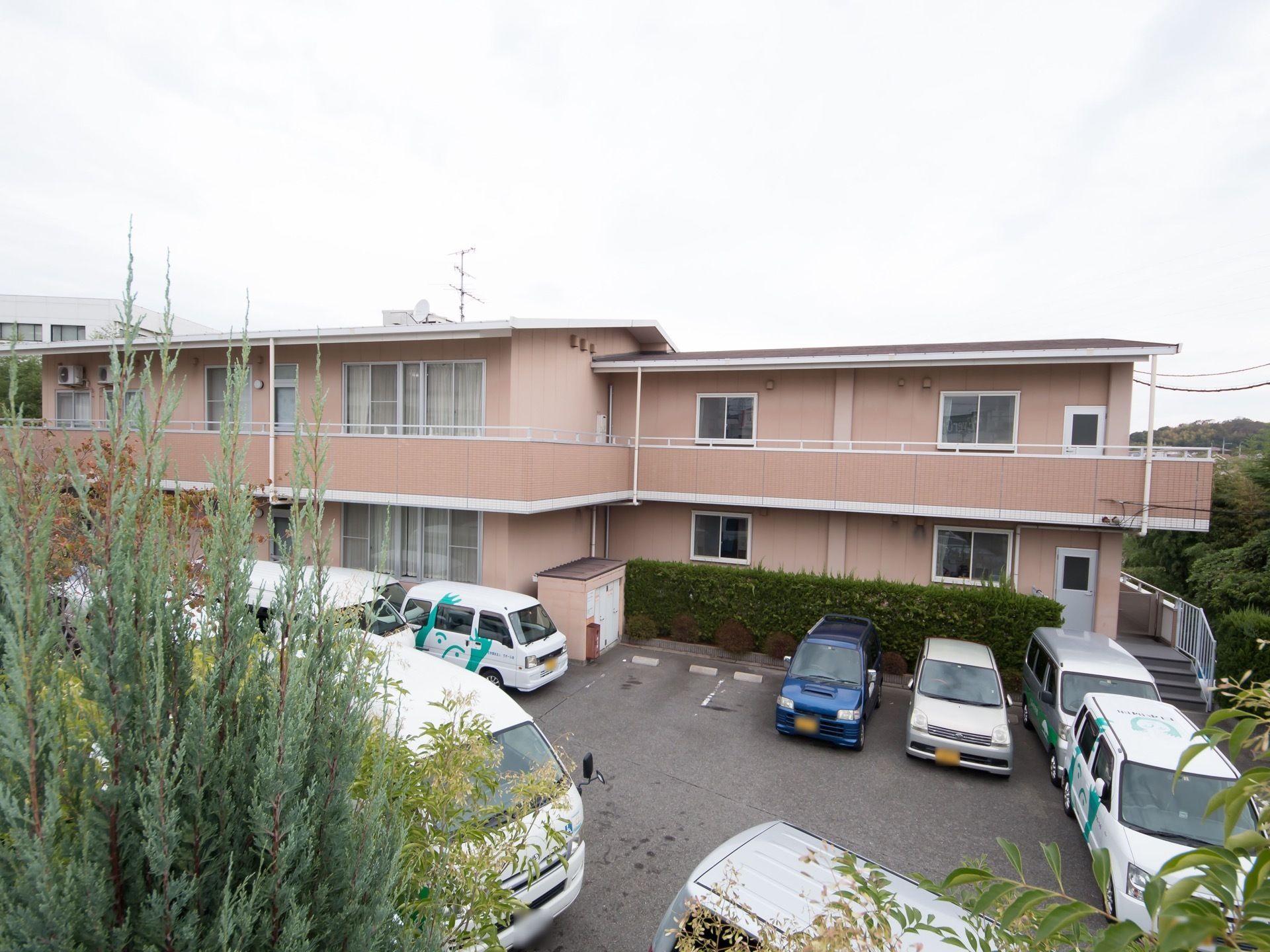 駐車場からみたでベージュの壁面の建物外観である。建物は斜面に建っているので、2階、3階部分が見える。