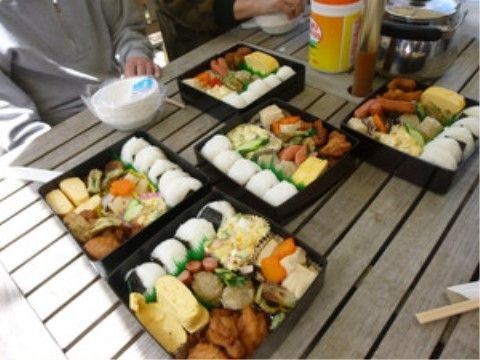 晴れた日の中庭で、木のテーブルの上に手作りのおにぎりや、おかずが美味しそうに詰められた重箱が並べられている。