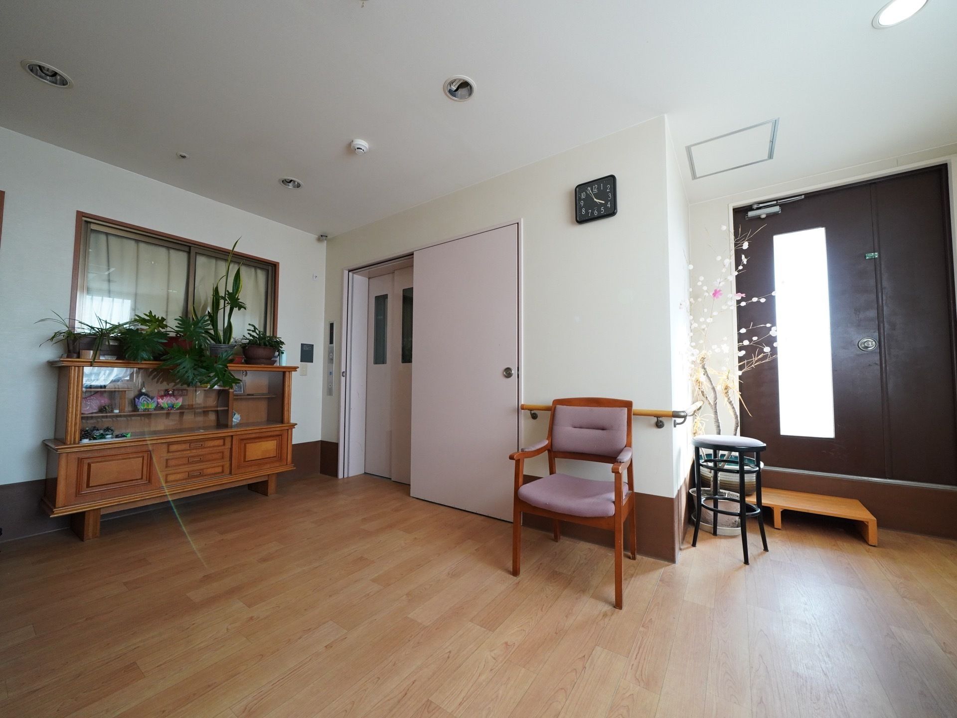 椅子が２脚置かれており、壁には時計が掛けられている。エレベーター前には棚があり、季節の飾りものや観葉植物が置かれている。