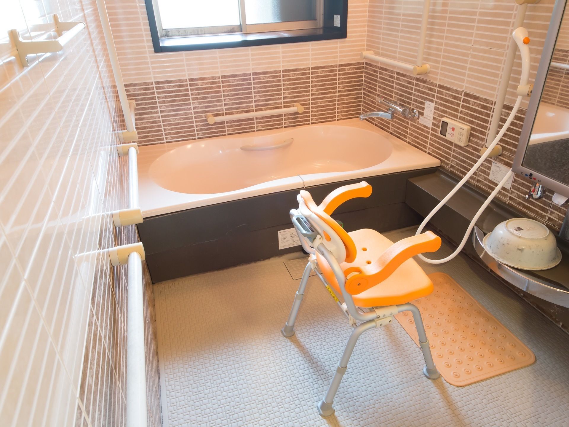 浴室はブラウンを基調とした落ち着いた雰囲気である。オレンジ色のシャワーチェアが置かれていて、床には滑り止めのマットがある。