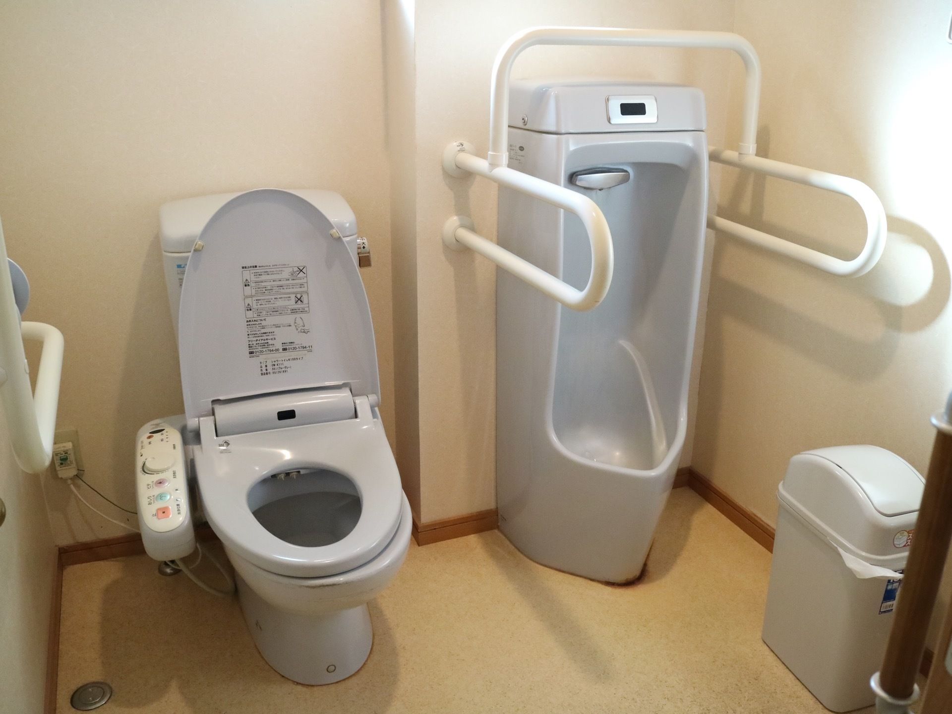 個室トイレの図である。薄いグレーの便座が２台並んでいる。左手には洋式トイレがあり、その横に男性用の便器がある。