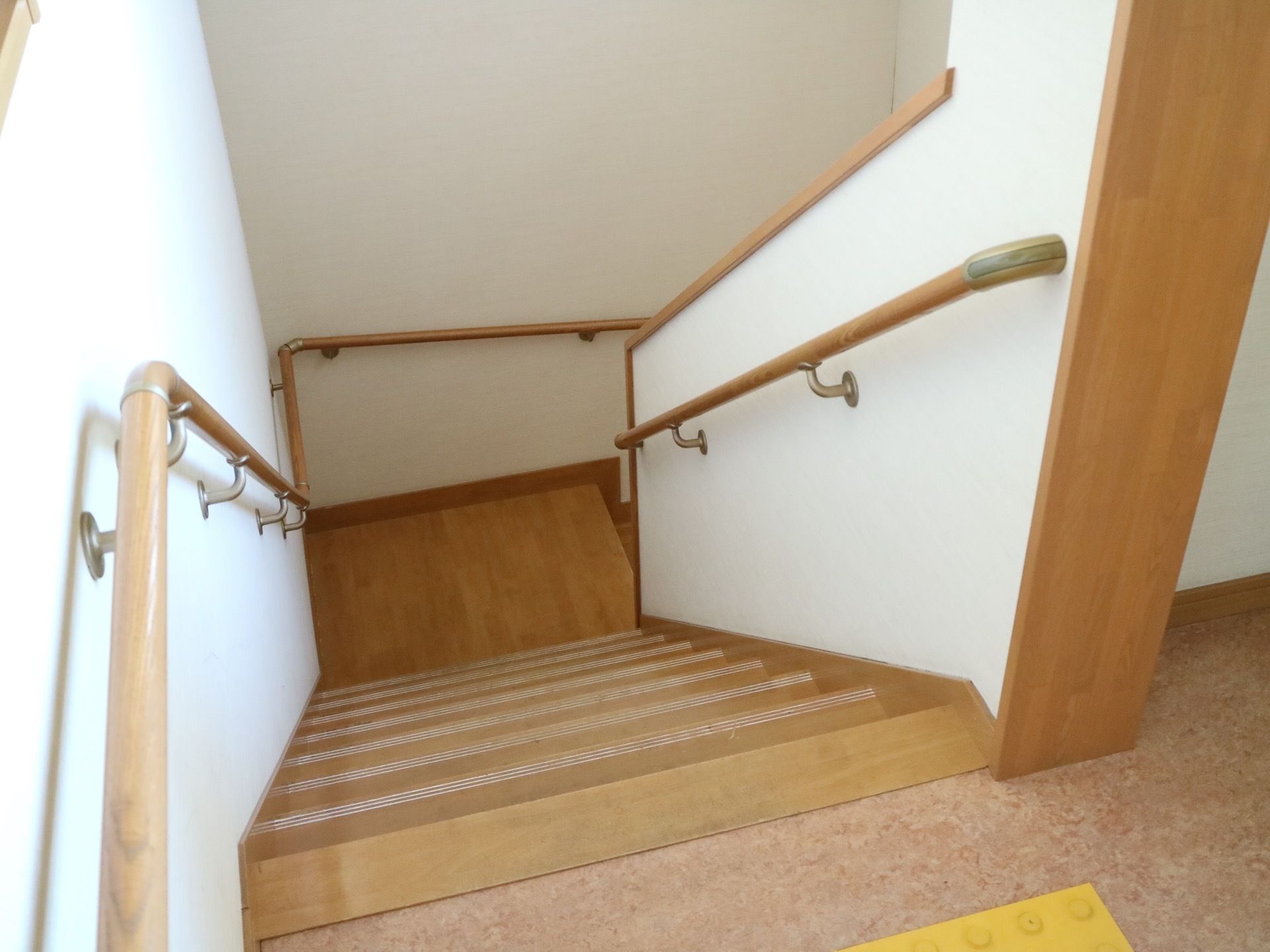 階下へ降りる階段の図である。白い壁に木製の手すりが両側に取り付けられている。床に視覚障碍者用のタイルが貼られている。