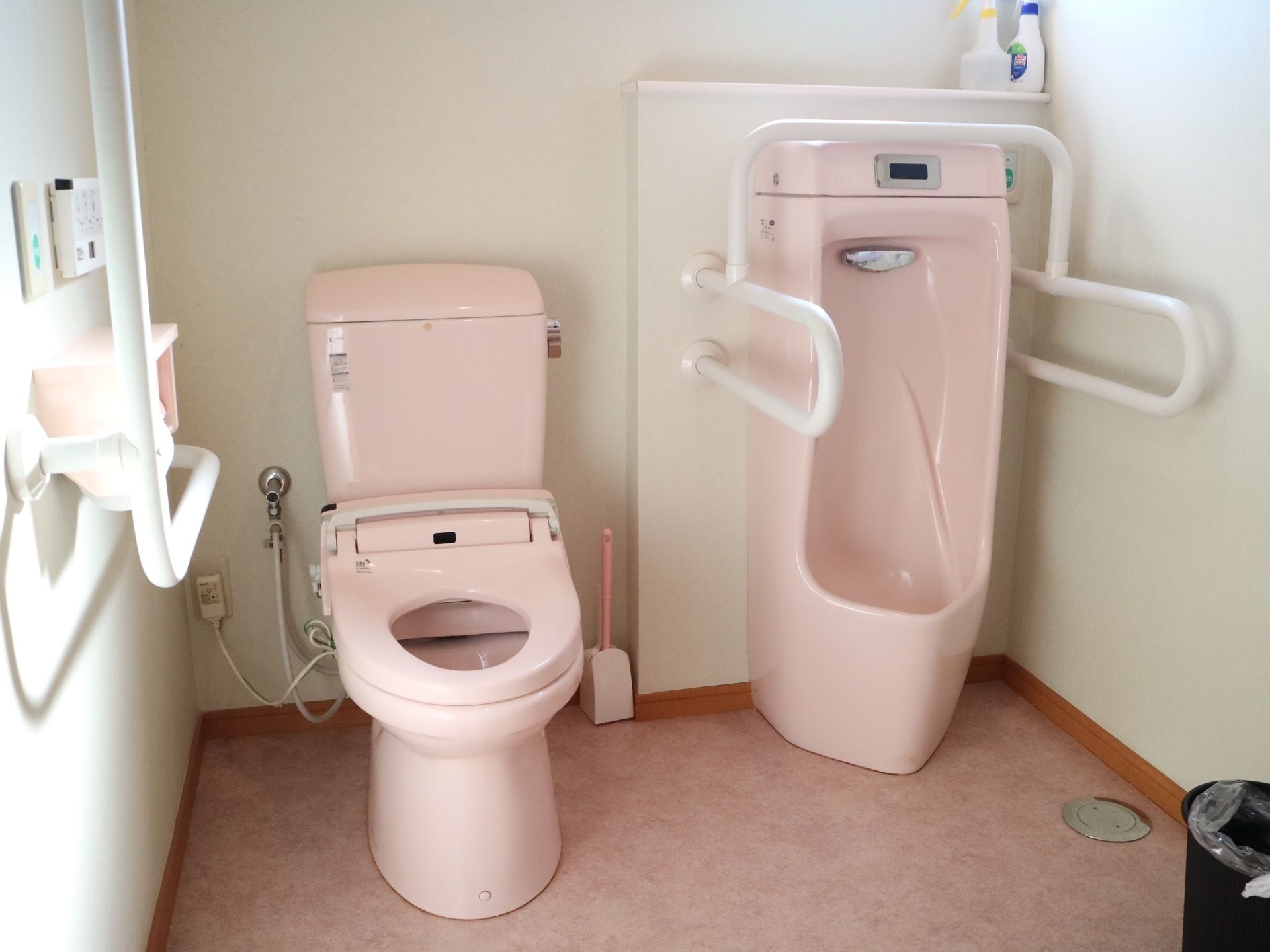 ２種類のピンクの便器があるトイレである。壁には白色の手すりが取り付けられており、小便器の前の棚には洗剤が置かれている。