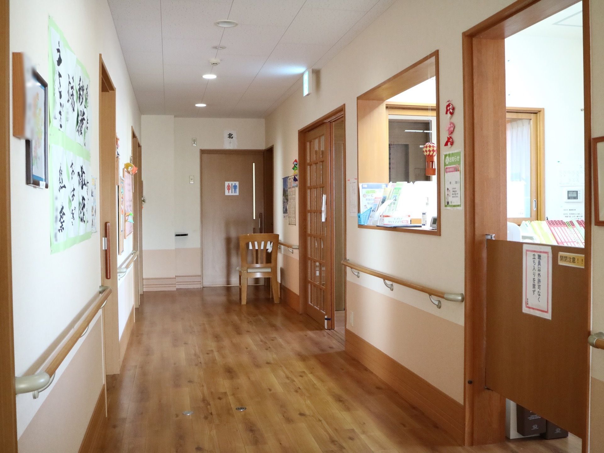 奥にはトイレの入口扉があり扉の前に椅子が置かれている。また、通路の白い壁には作品や絵が展示されている。