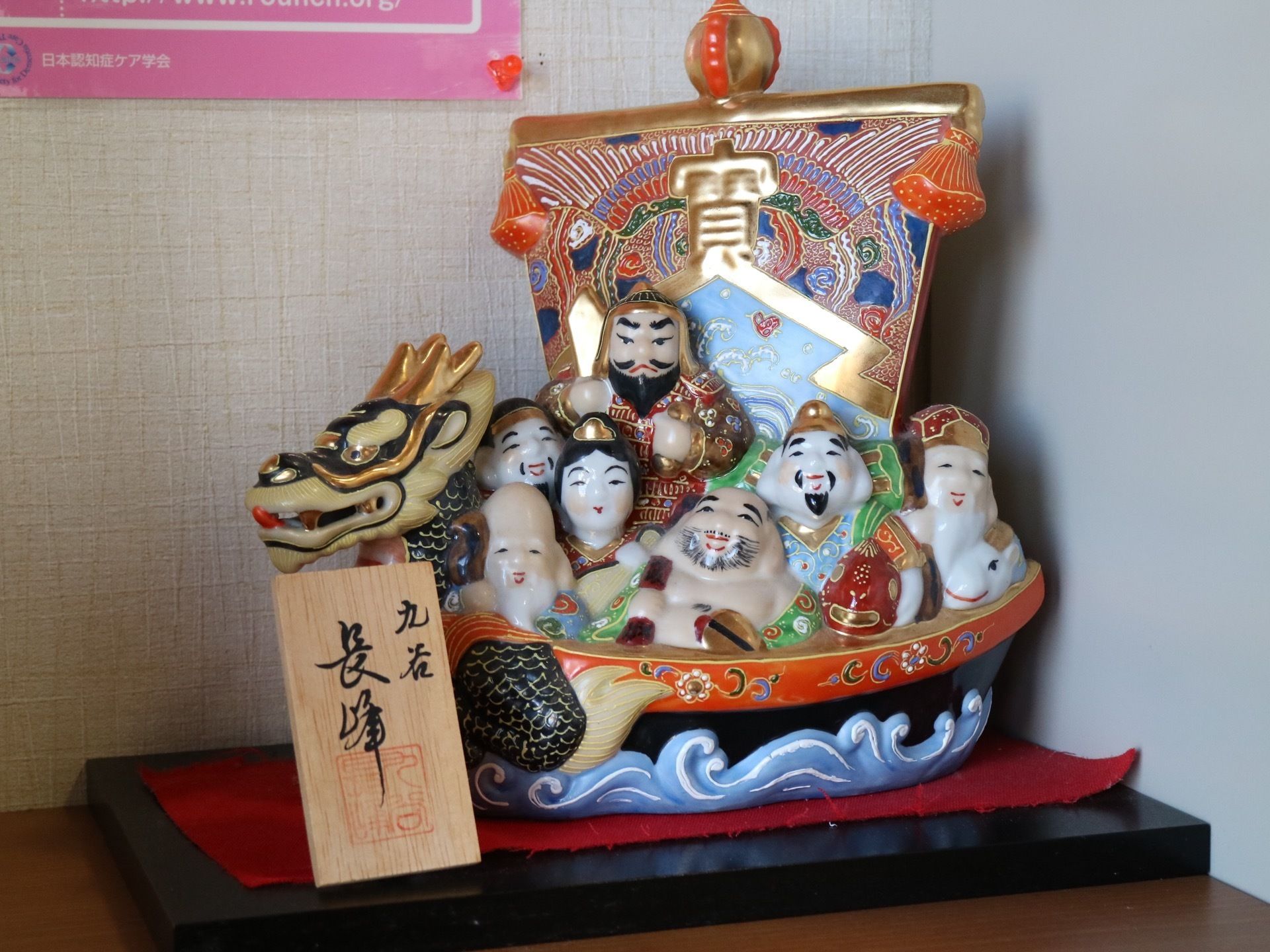 茶色の棚板の上に、黒色の台があり、その上に七福神の置物が置かれている。置物は九谷焼である。後ろの壁には、掲示物が貼られている。