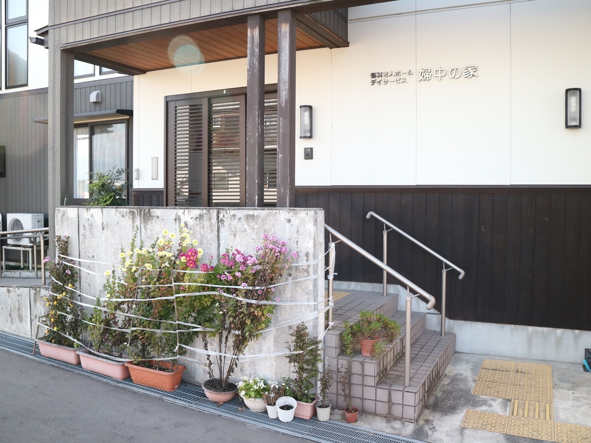 白い壁の間に黒い格子の玄関扉がある。玄関前には、複数のプランターが並べられており、植物が植えられている。