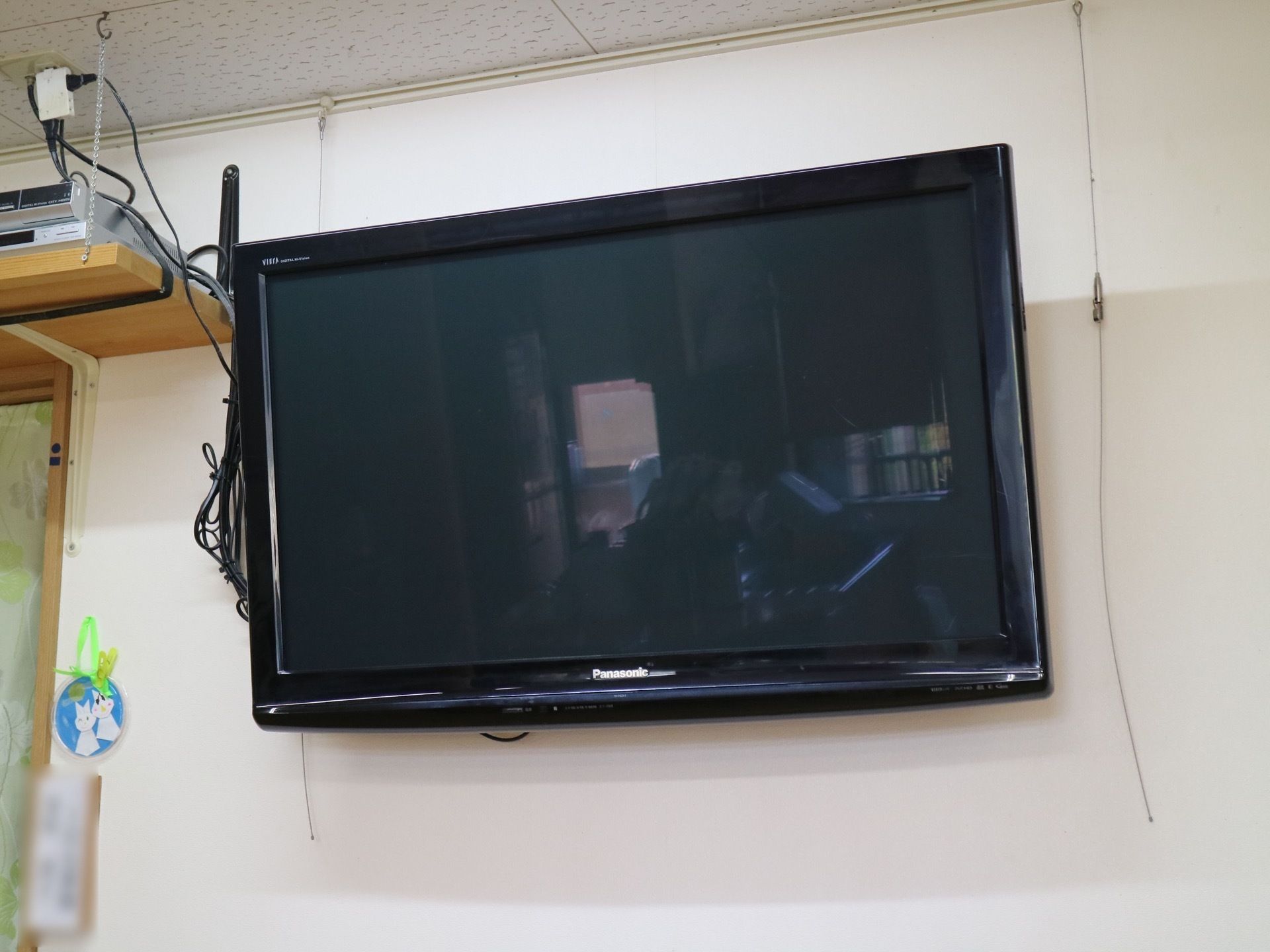 白い壁の中央に大型液晶テレビが取り付けられている。左側の壁の天井近くに木材でできた棚が１段あり、コンセントがつながっている。