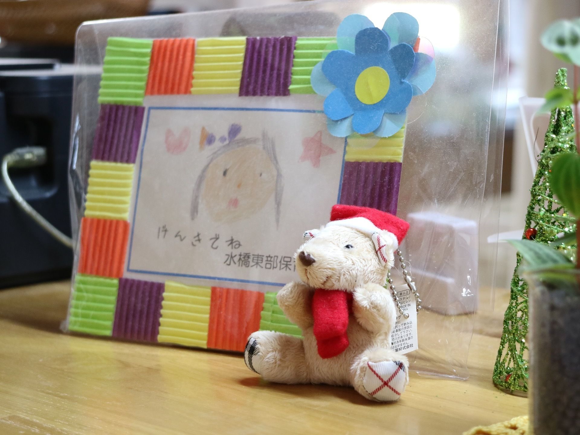 木製の棚の上に置かれた写真立ての図である。手書きの似顔絵が書かれたものが入れられている。熊の人形がある。