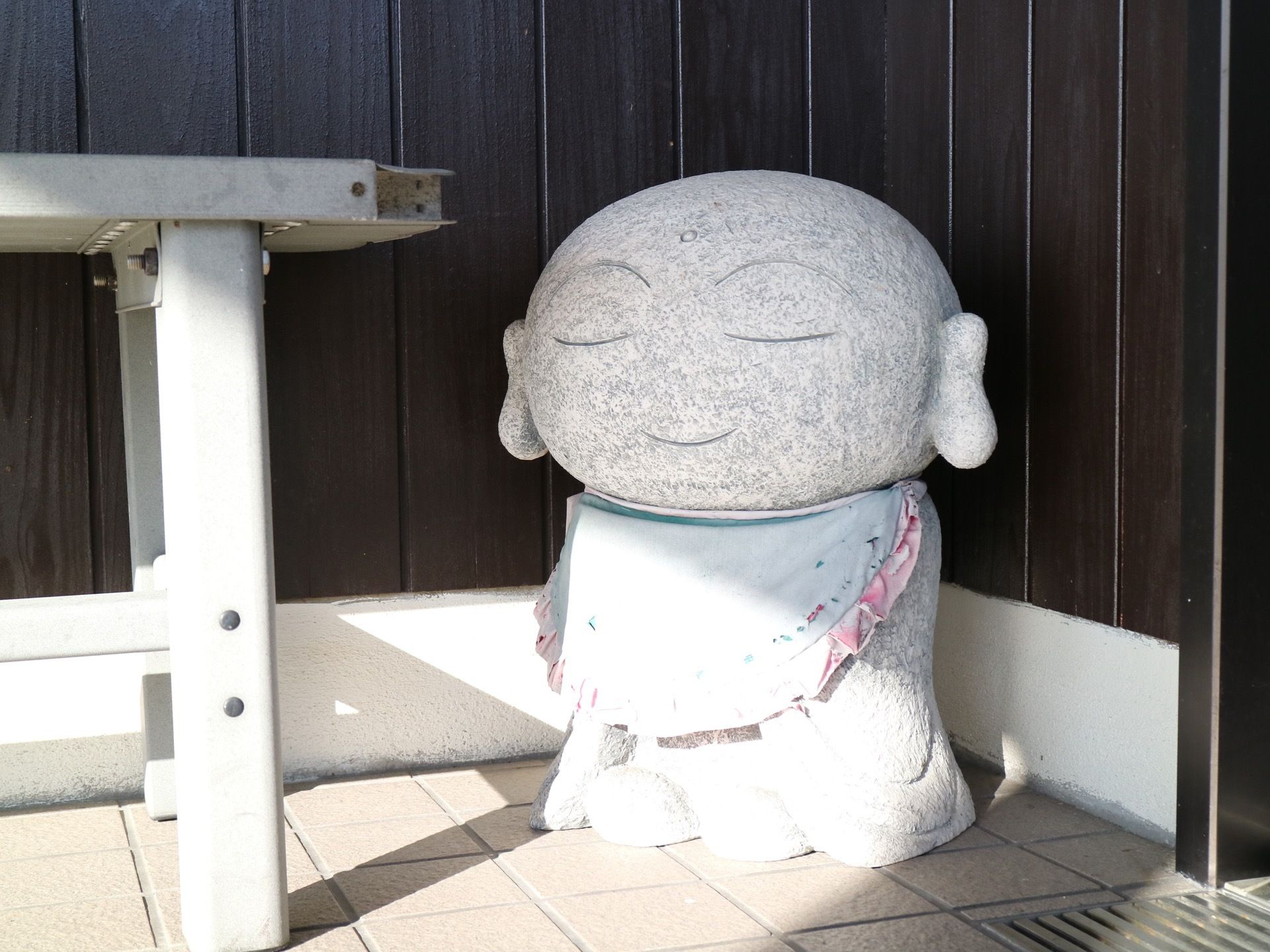 茶色で縦長の木材が貼られた外壁に、石畳の施設外部である。中央に石でできた大きなお地蔵様が置かれている。