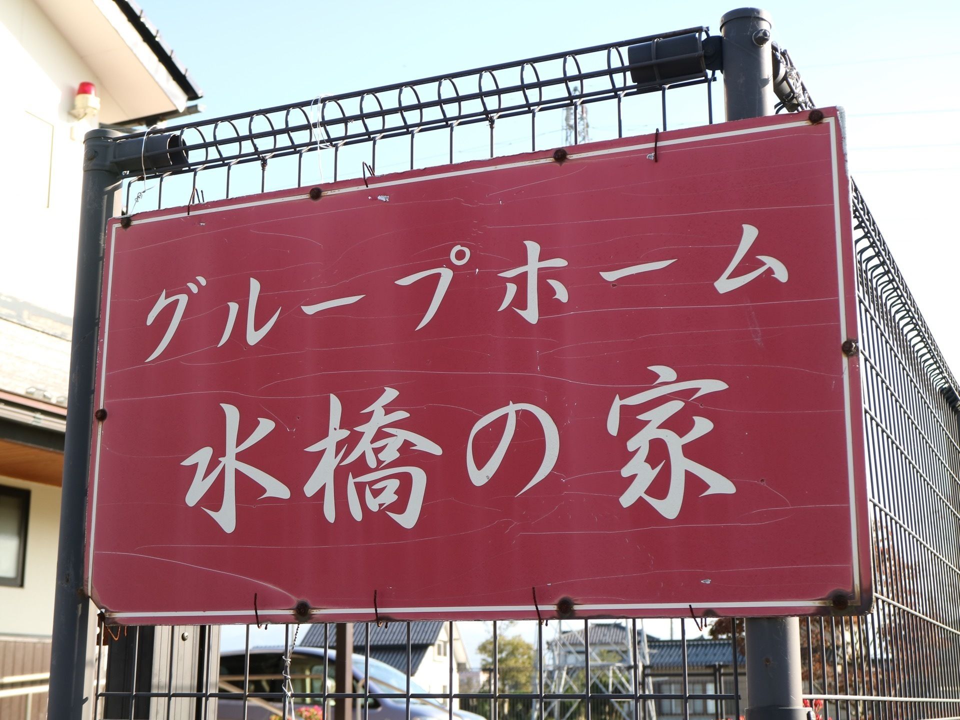 施設の外に設置された看板の図である。茶色の金網に、白い字で施設名が書かれた赤い大きな板が取り付けられている。