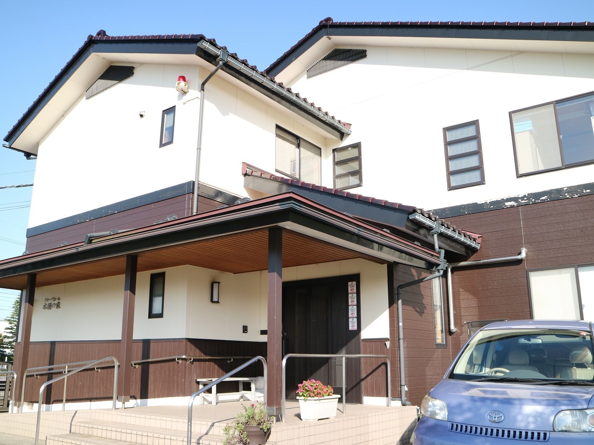 一軒家のような佇まいの施設で、白い外壁に黒い瓦屋根がのっている。施設の前に自動車が１台停まっている。