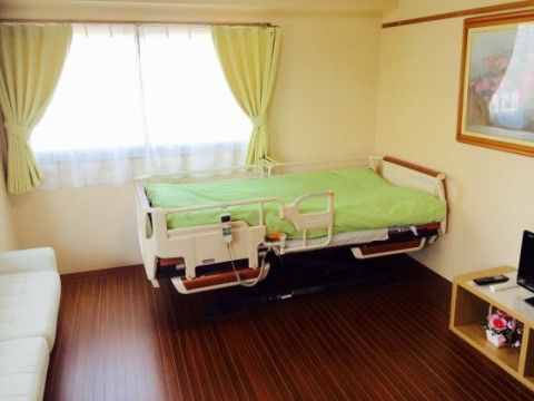 床は濃い茶色のフローリングで、壁はベージュ色で落ち着く印象の居室である。布団のシーツやカーテンが緑色である。