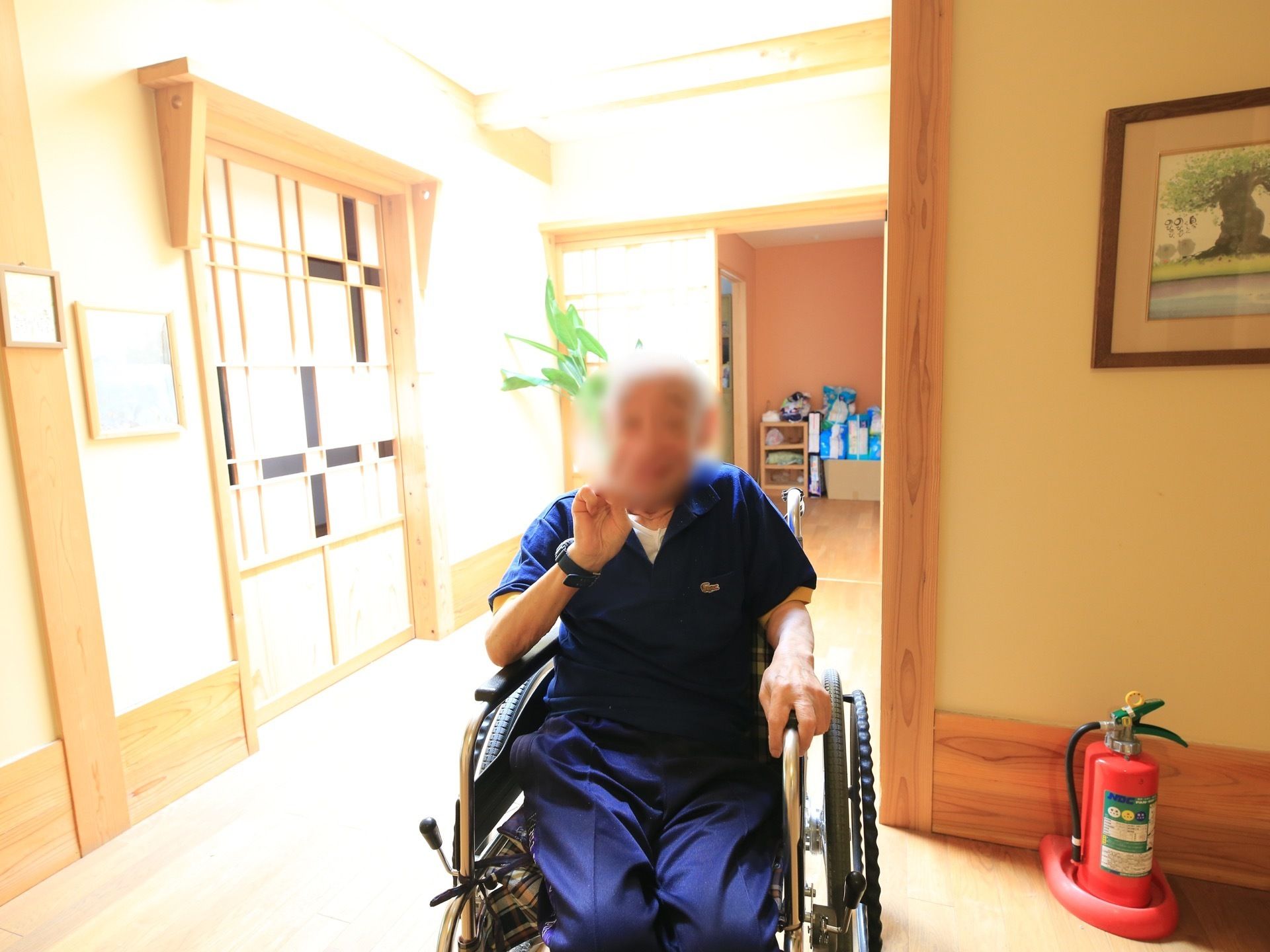 居室前の日当たりの良い廊下で撮影された写真である。車椅子に座った男性の入居者様が、笑顔でピースしている。