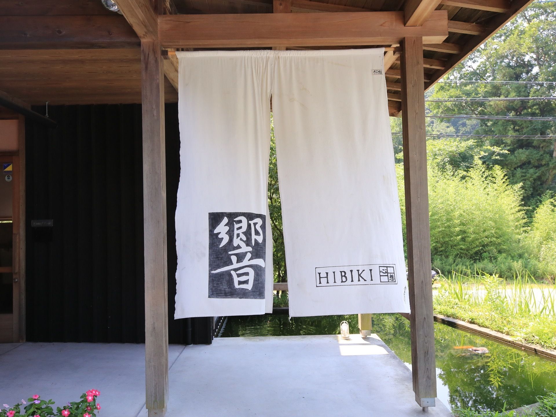 施設玄関に、施設名が書かれた白いのれんが掛けられている。横には、鯉が泳ぐＬ字型の中池がある。