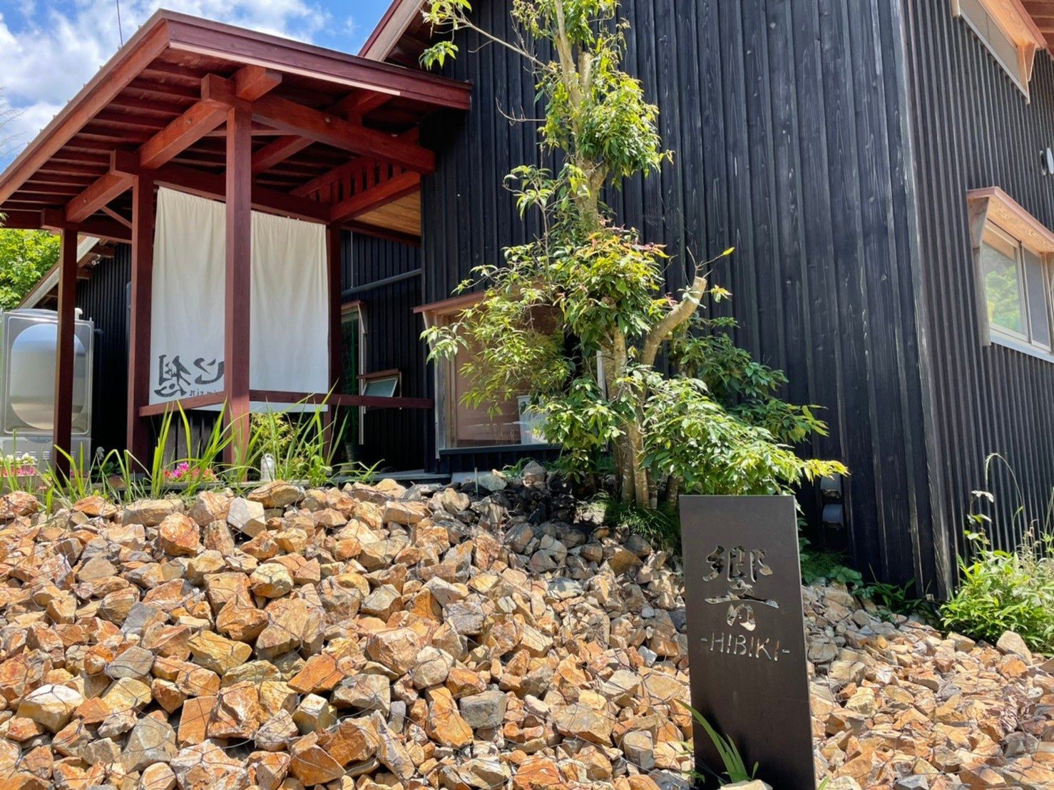 木造の平屋建てのグループホームで、周辺は緑豊かな山々に囲まれている。手前には車が１台停められている。