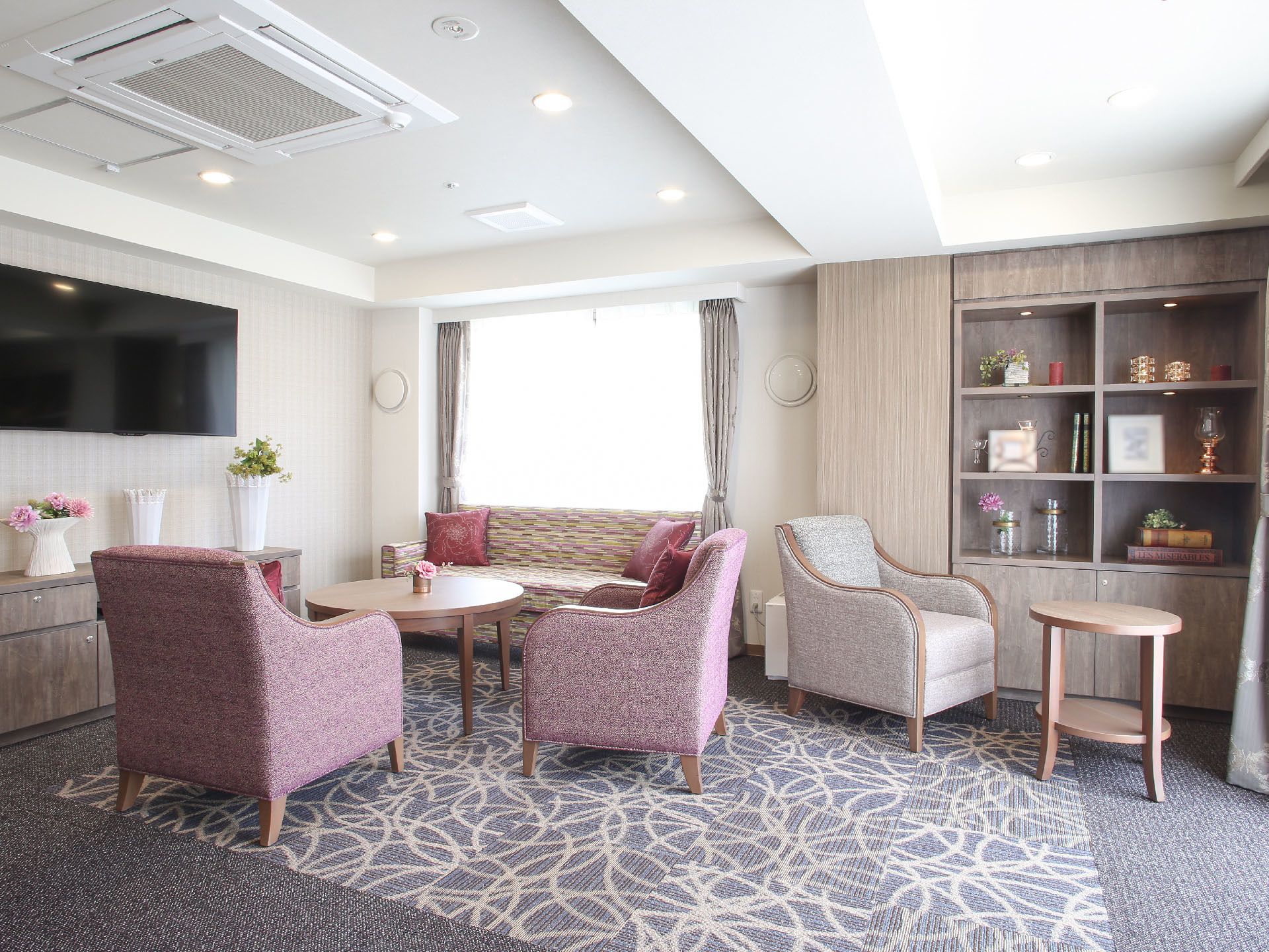 壁掛けの大きなテレビが設置された部屋である。丸型テーブルやゆったり座れるソファ、収納棚が体裁よく配置されている。