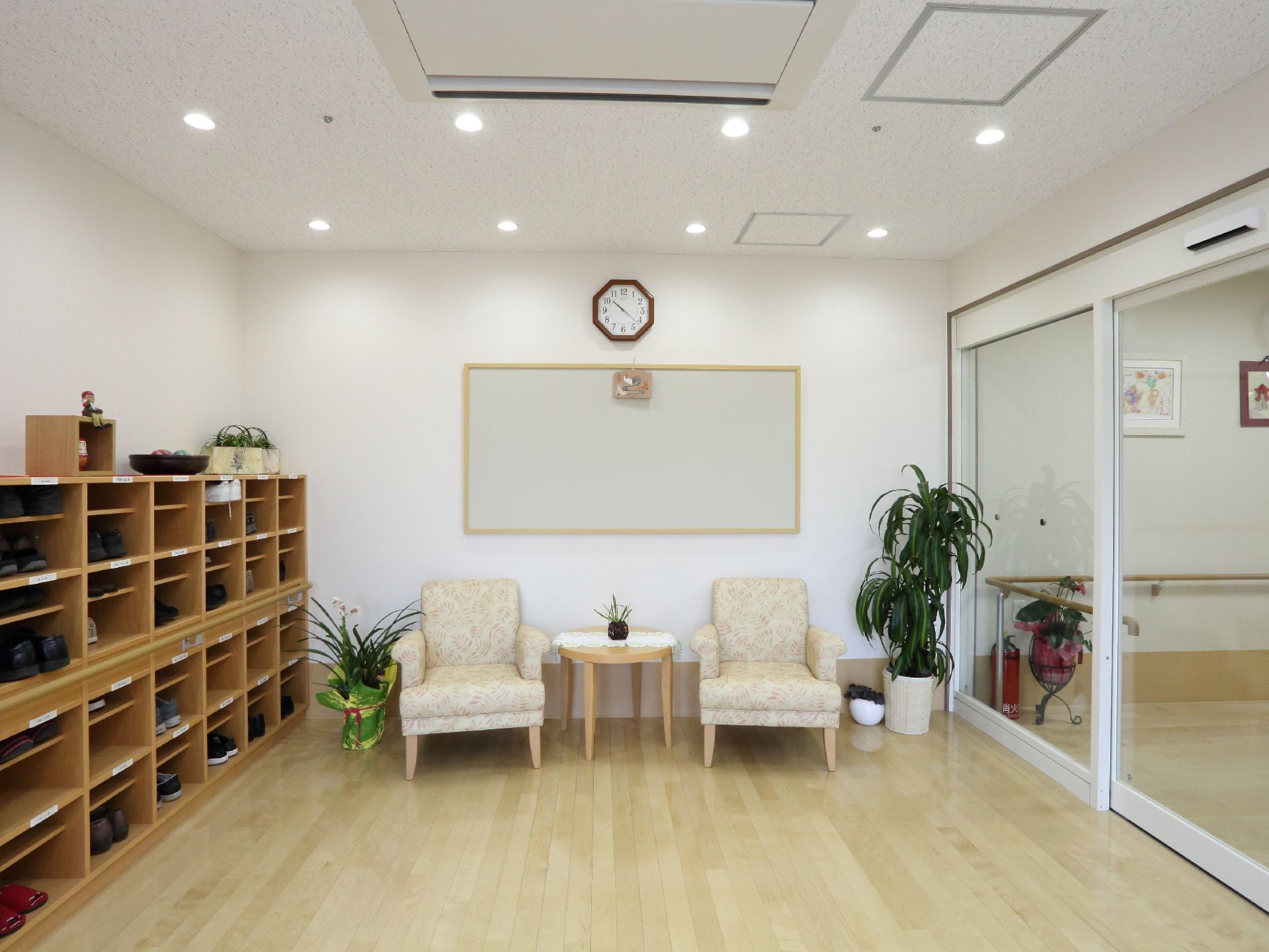 真ん中には椅子が二脚置かれている。それを囲うように植物が置いてある。左には靴箱があり壁にはボードが設置してある。