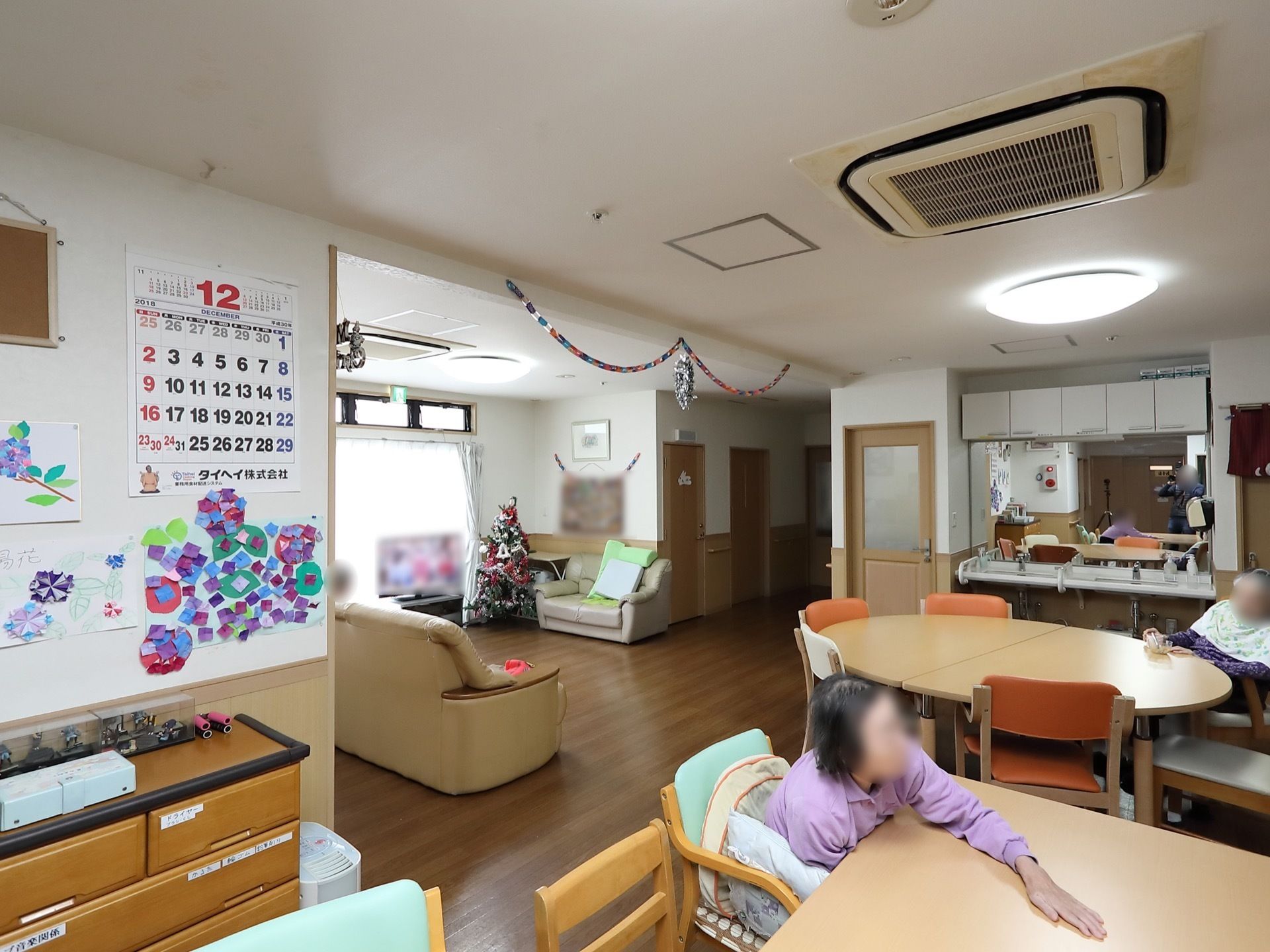 広いスペースに丸や四角いテーブル、ソファなどが置かれている。壁にはカレンダーや、折り紙で作った花の絵などが飾られている。