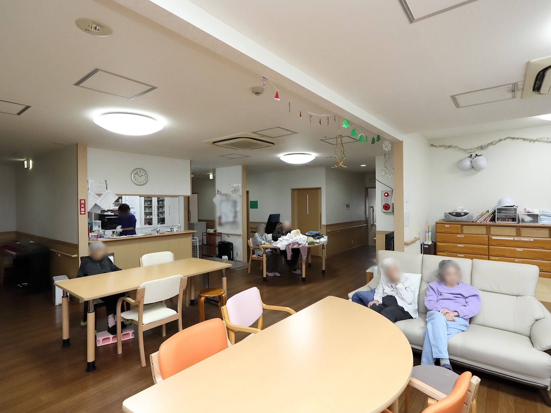 テーブルや椅子、ソファなどが用意され、入居者様が座っている。奥にはキッチンがあり、スタッフが作業をしている。