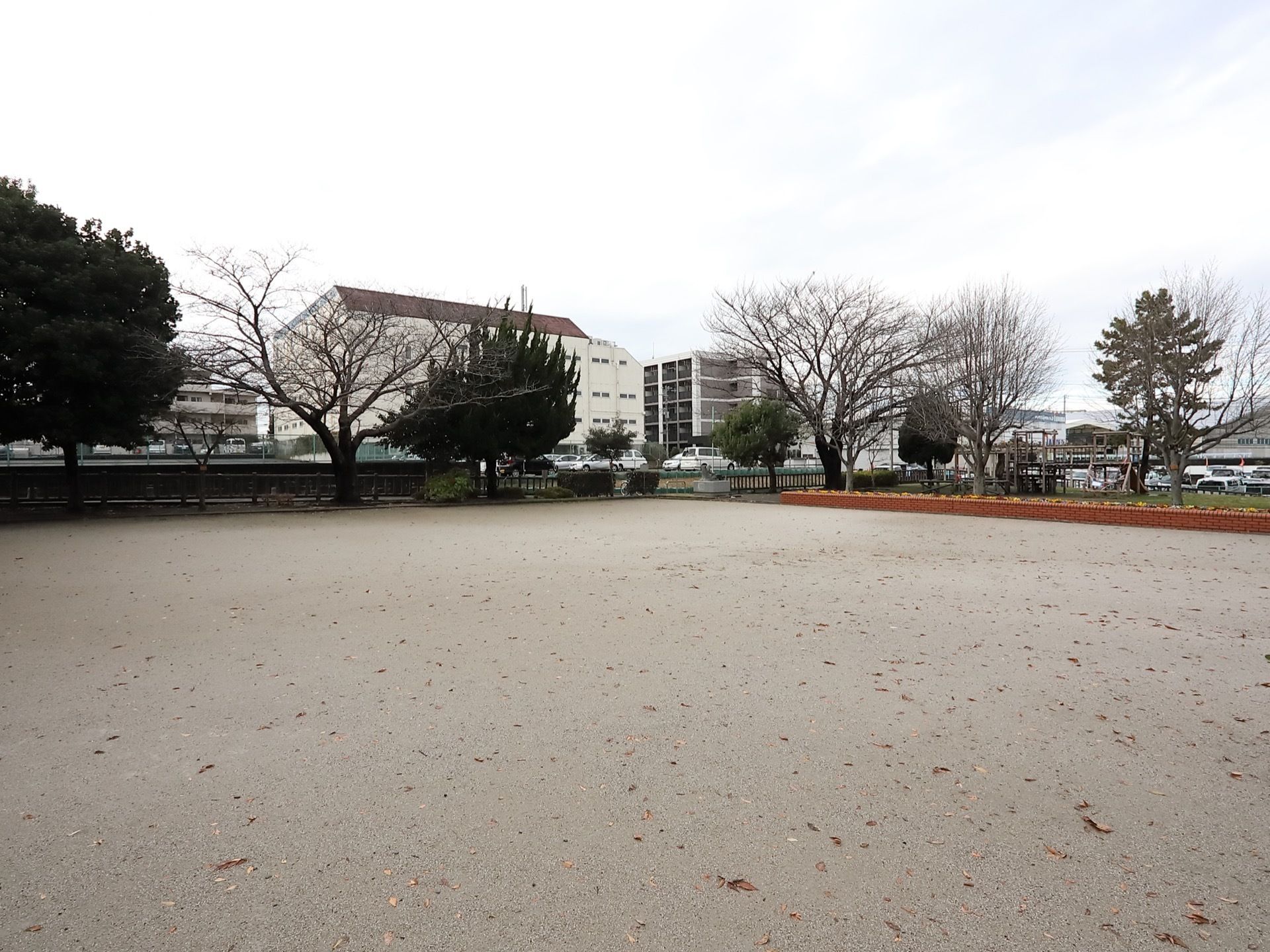広々とした公園で、周りには木がたくさん植えられている。奥の方には大きな建物があり、その手前には車が数台停められている。