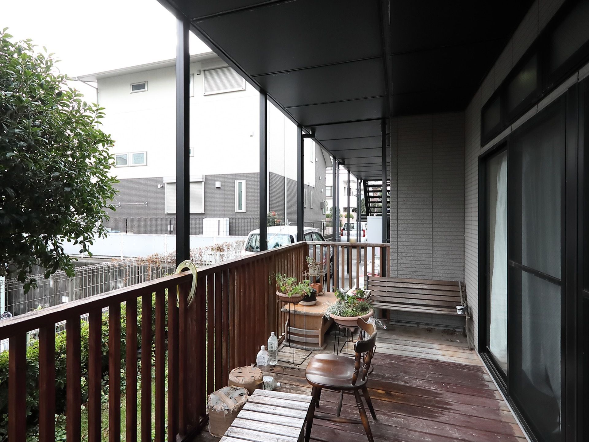 ウッドデッキには椅子やベンチ、鉢植えなどが置かれている。上には黒い屋根がついており、外には木が植えられている。