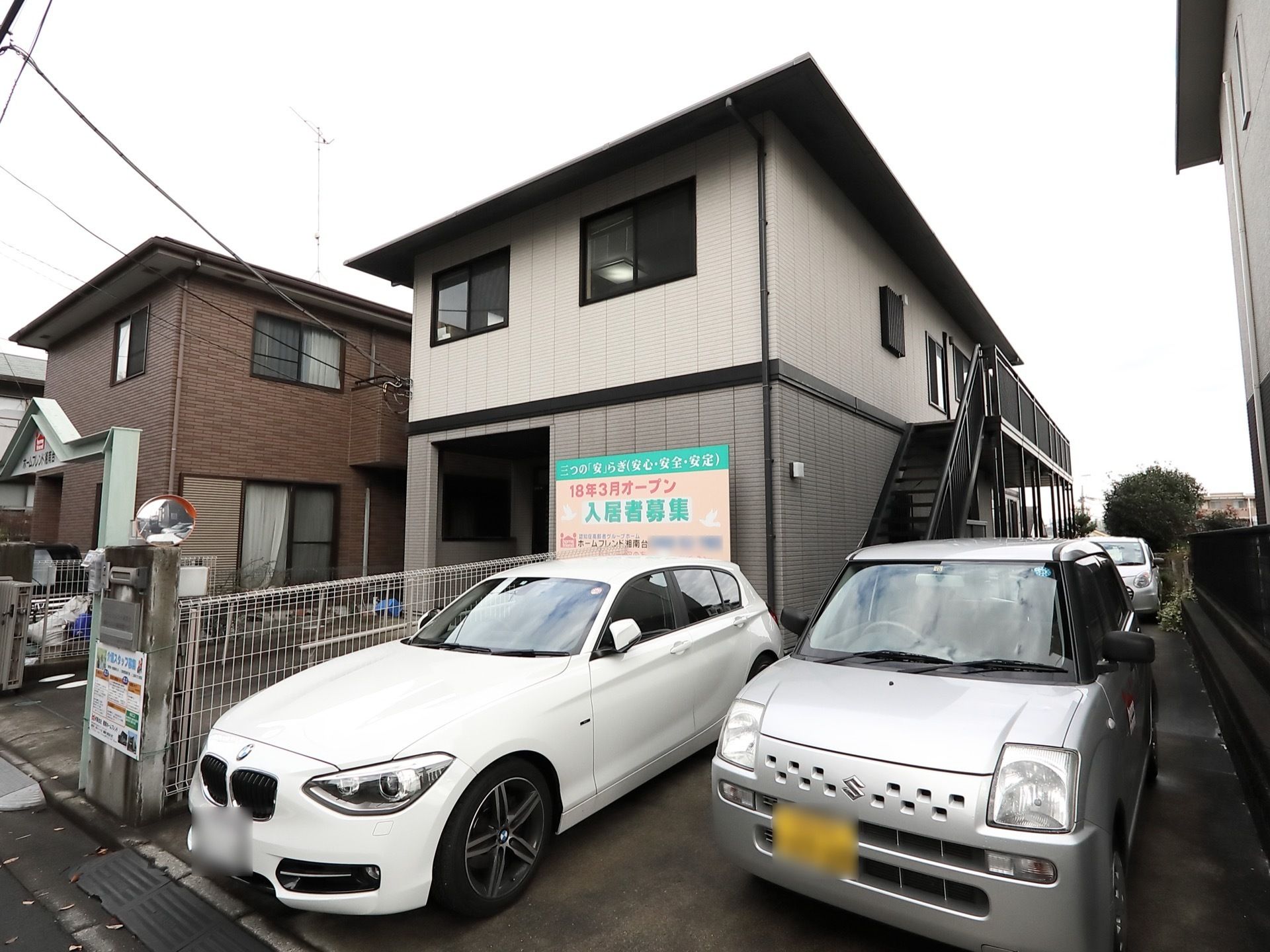 ライトグレーの２階建てのホームである。入り口のすぐ横が駐車スペースになっており、２台の車が停まっている。その奥には外階段がある。