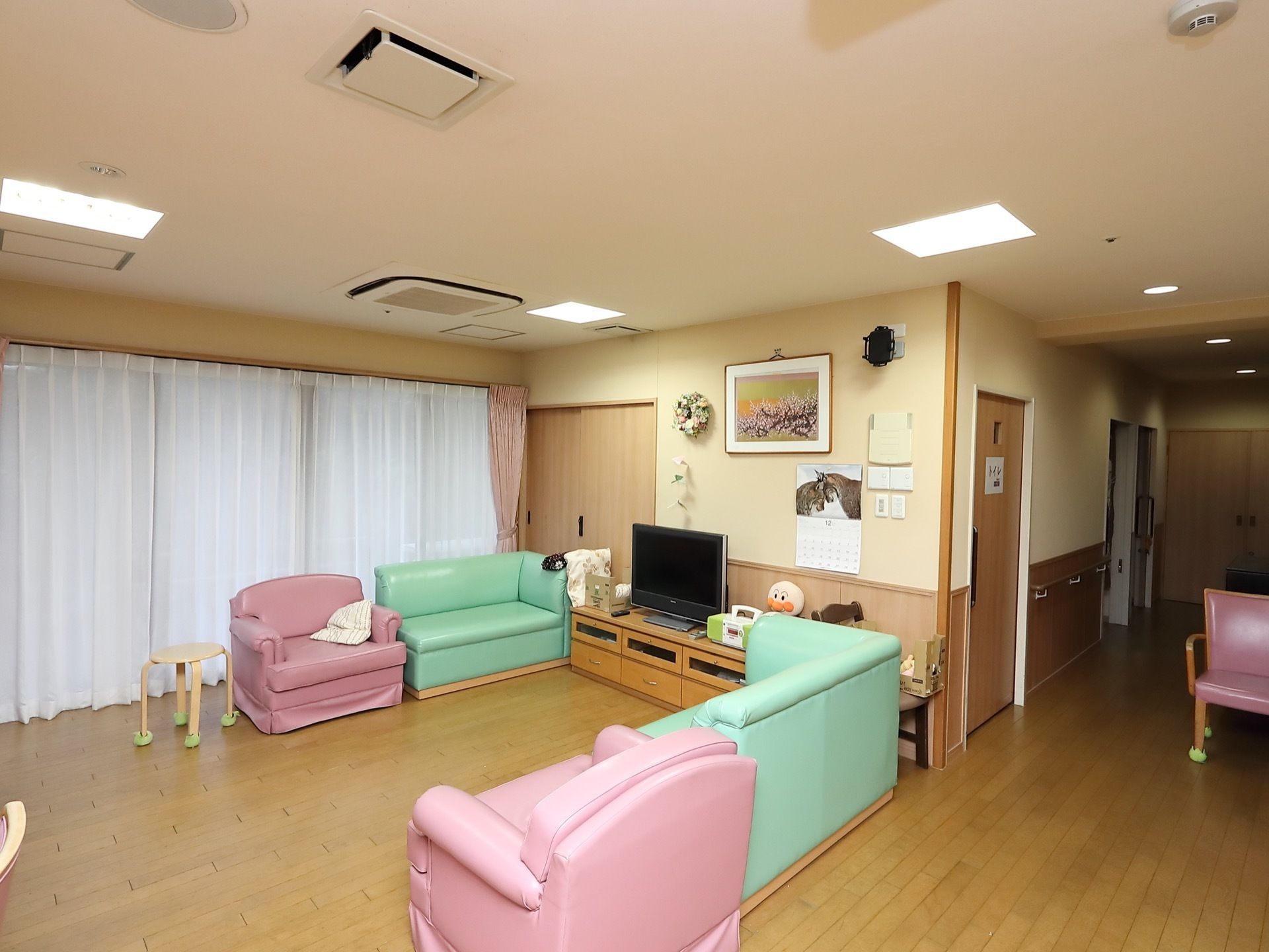 ピンクとグリーンのソファが向かい合わせに置かれている。壁際にはテレビなどがあり、壁には絵やカレンダーが飾られている。