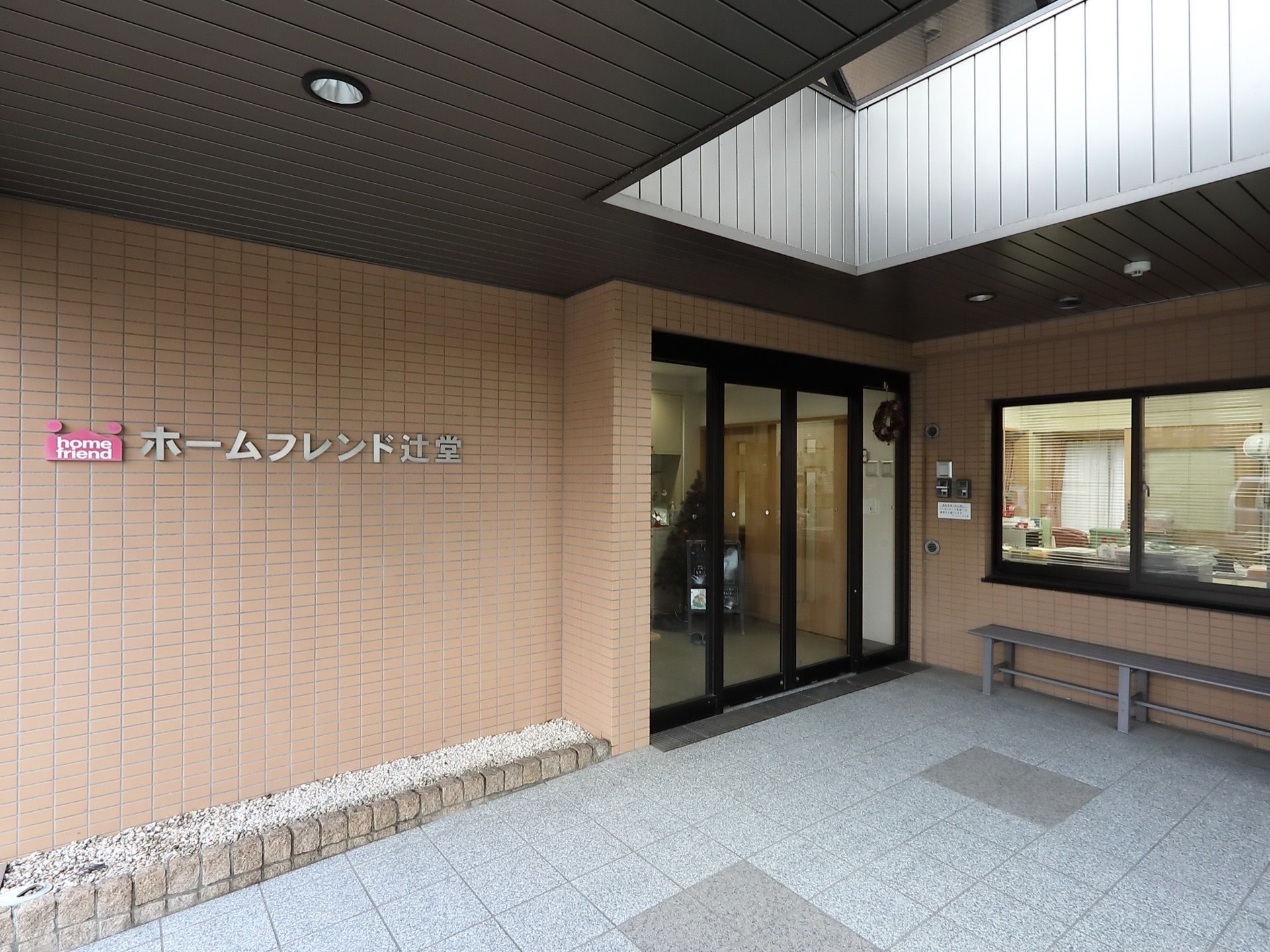 ベージュのレンガ調の壁に、施設名が書かれている。出入り口は自動ドアになっており、入口の前にはベンチが置かれている。