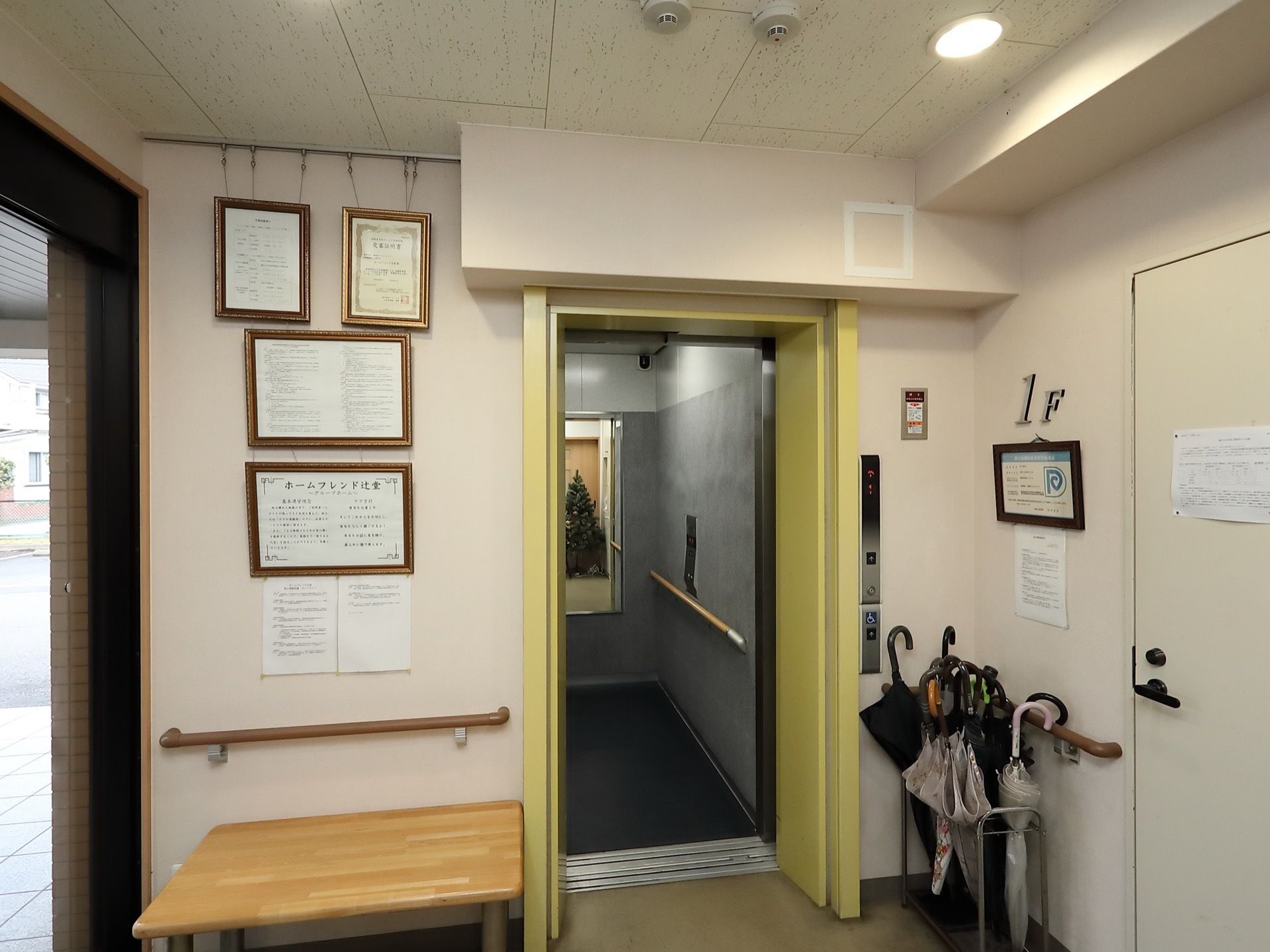 壁には手すりがついており、右の壁際には傘立てが置いてある。エレベーターのドアが空いており、中には鏡が設置されている。