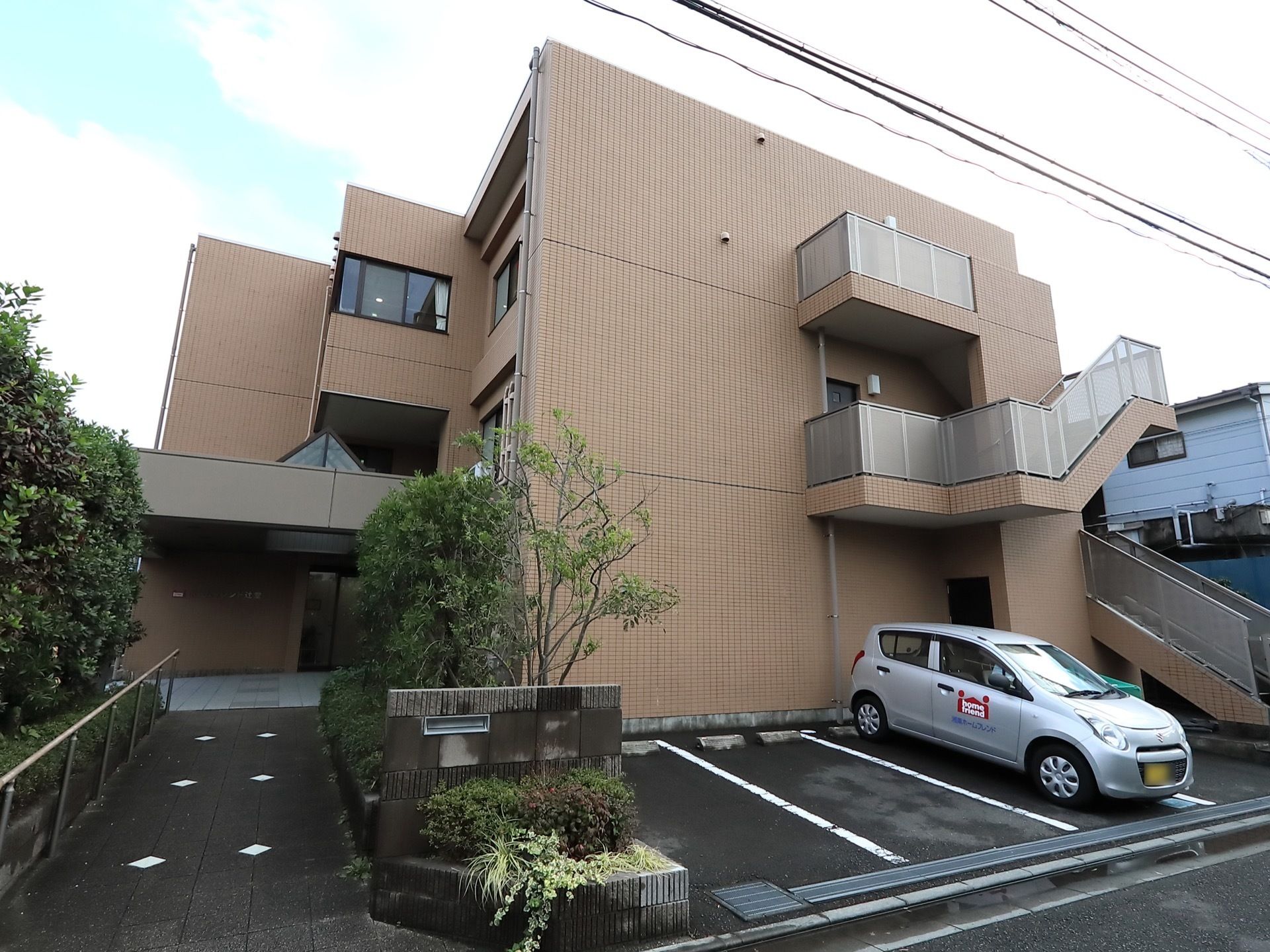 茶色い３階建ての建物で、右側には外階段がある。建物の前の左側はスロープになっていて、右側には駐車場がある。