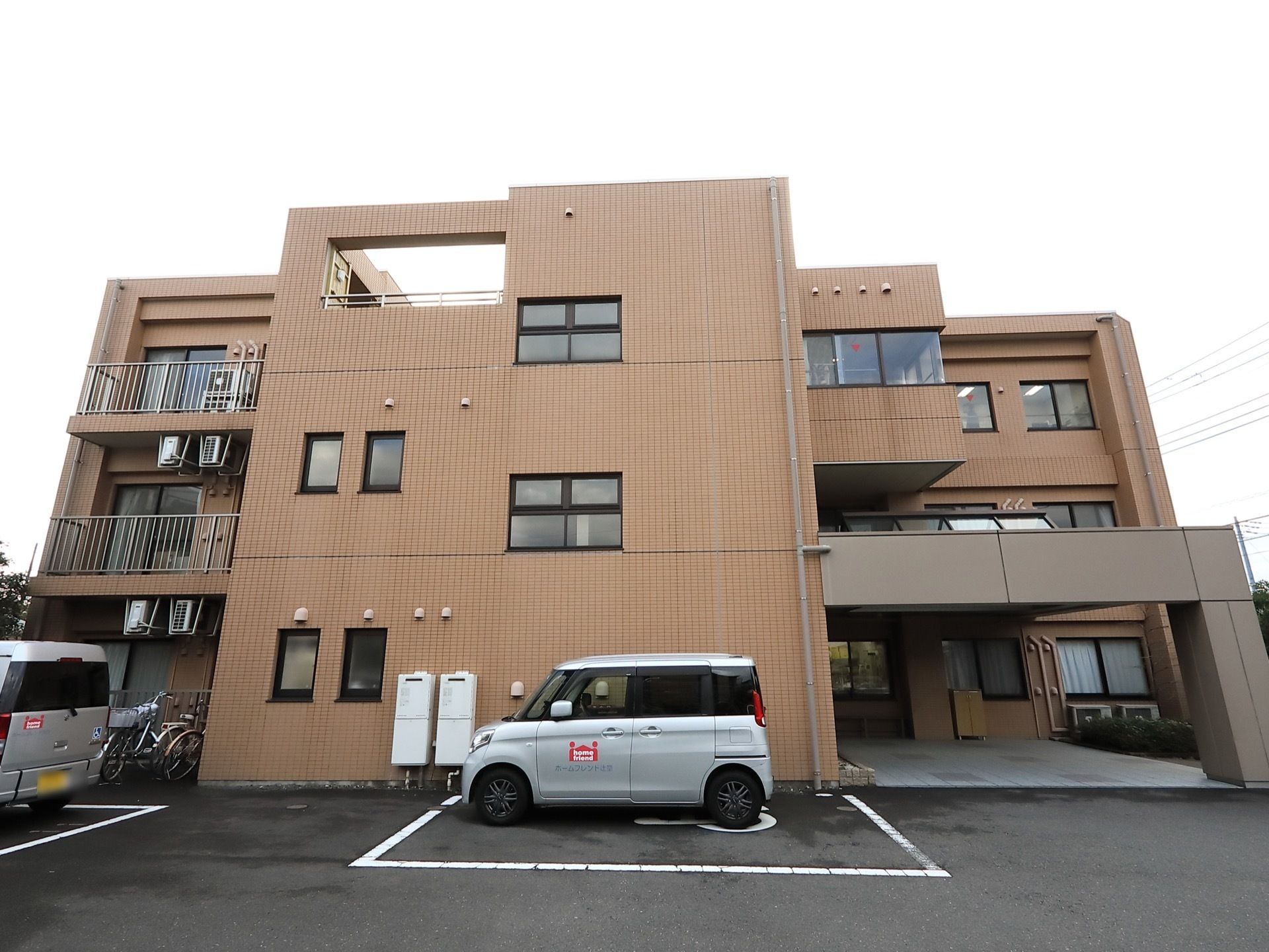 ３階建ての建物の左の部分にはベランダがついている。建物の前には車椅子マークが書かれた駐車スペースがあり、そこに1台車が停まっている。