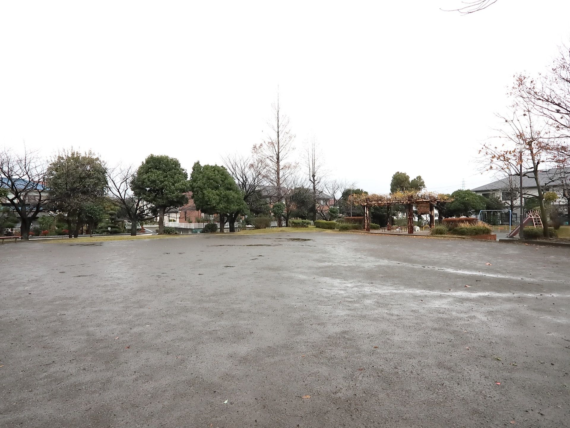 広場のある公園で、周囲には樹木が植えられていて藤棚が設置されている。片隅にはすべり台とブランコが見える。