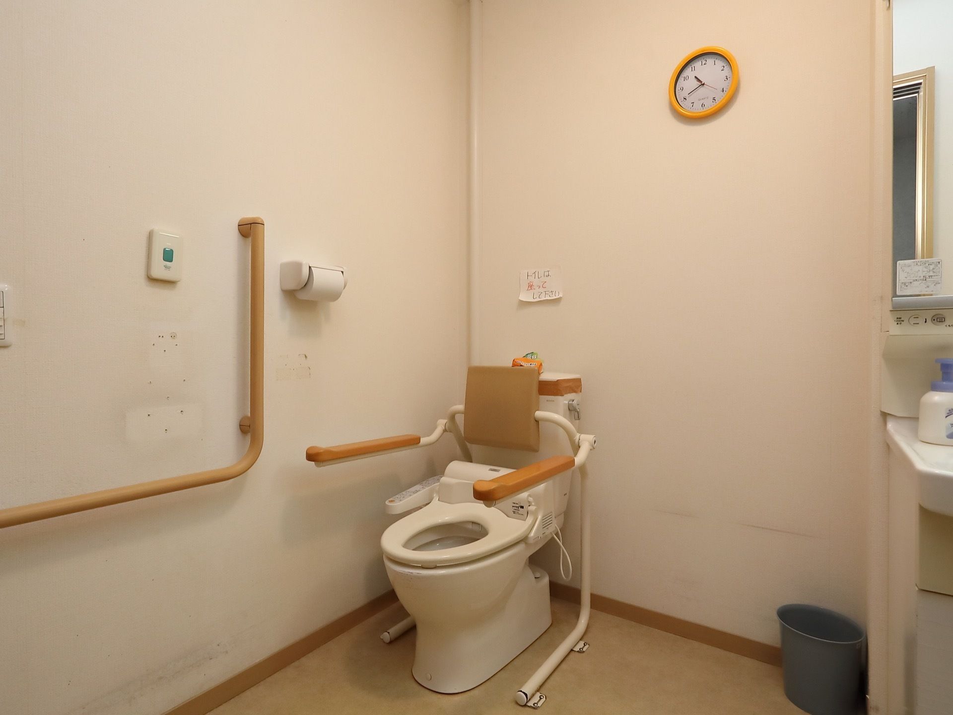洋式便器が一据ある。壁にはＬ字型の手すりが設置され、時計が掛けられている。隣には洗面化粧台が見える。
