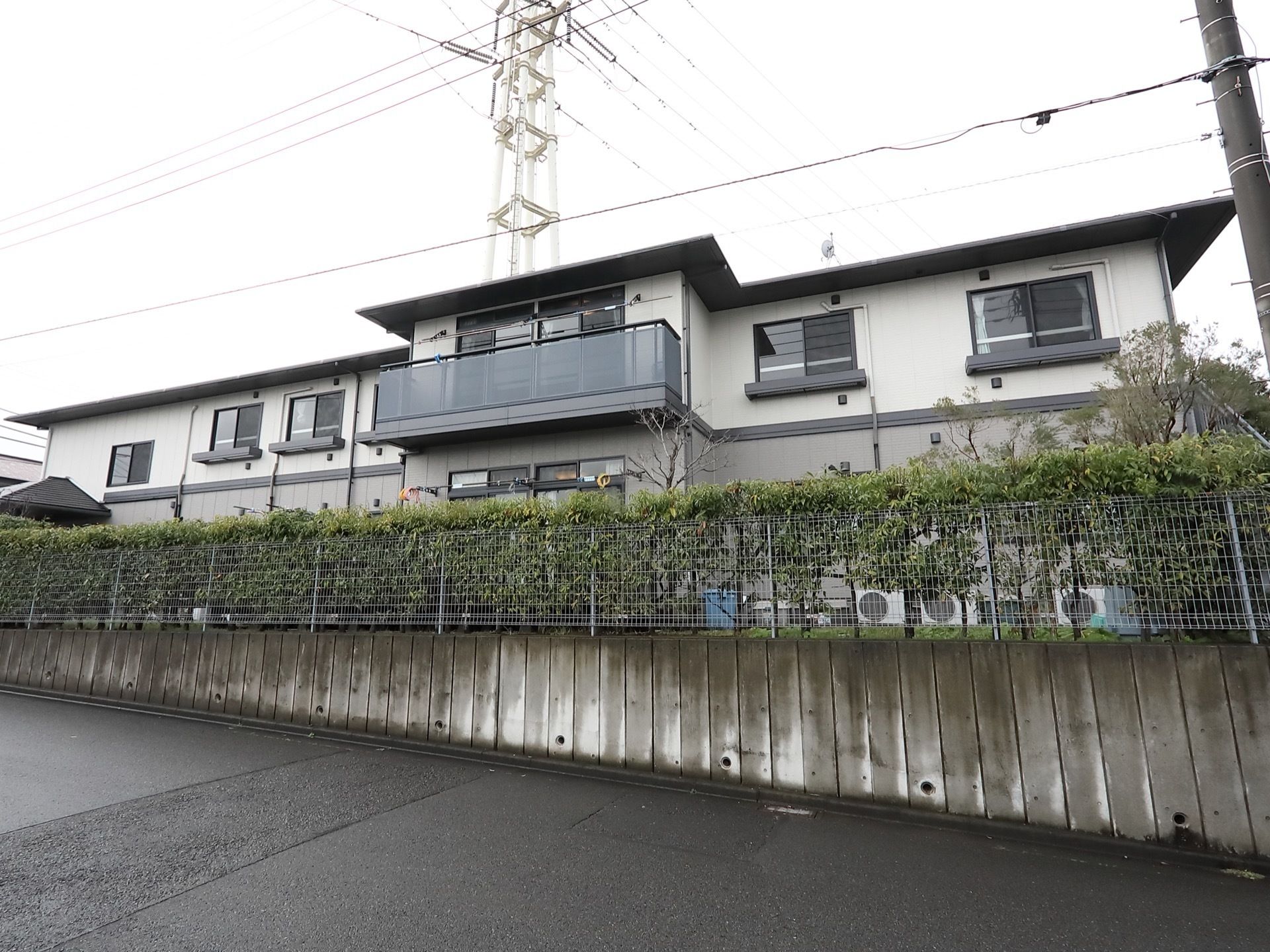 ２階建ての施設が見える。建物の中央にはバルコニーが設けられている。周りにはフェンスが設けられており、内側には木が植えらえている。