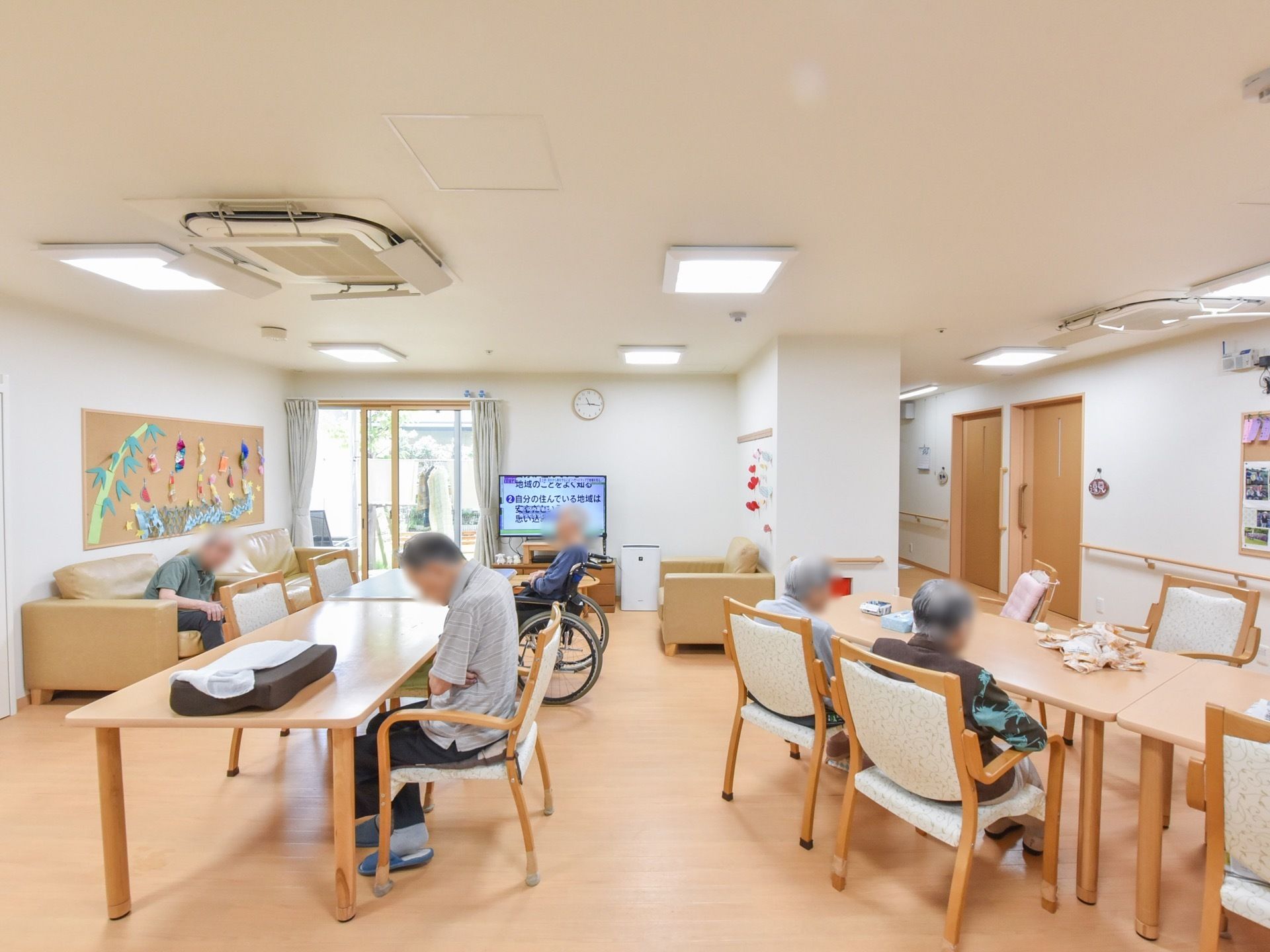明るい色調でやさしい印象の共有スペースには、テーブルセットやソファーが設置され、椅子に腰かける数名の男女の入居者様の姿が見える。
