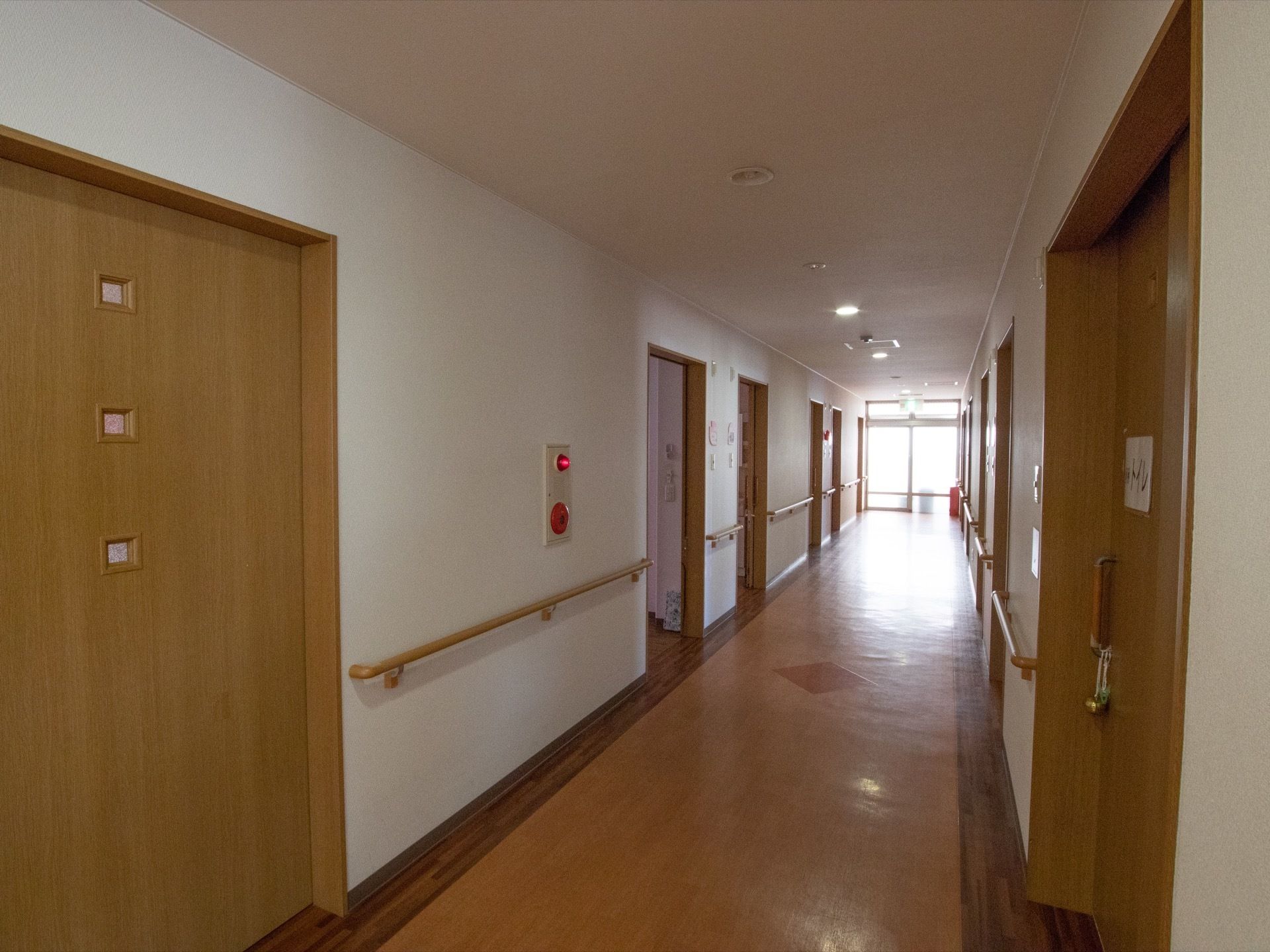 広い廊下に個室の扉が並んでいる。壁には手すりが設置されている。突き当りに大きな窓があり、日差しが入ってきて明るい。