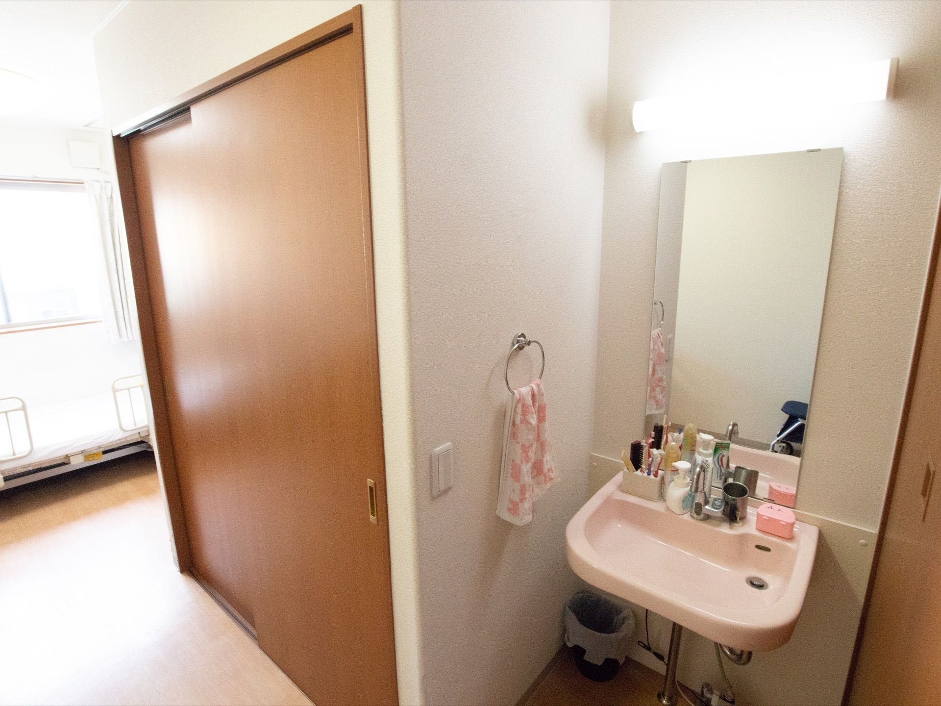 大きな鏡とピンク色のシンプルな洗面台があり、鏡の上部には蛍光灯、壁にはタオル掛けがある。コップや歯ブラシを置くスペースがある。