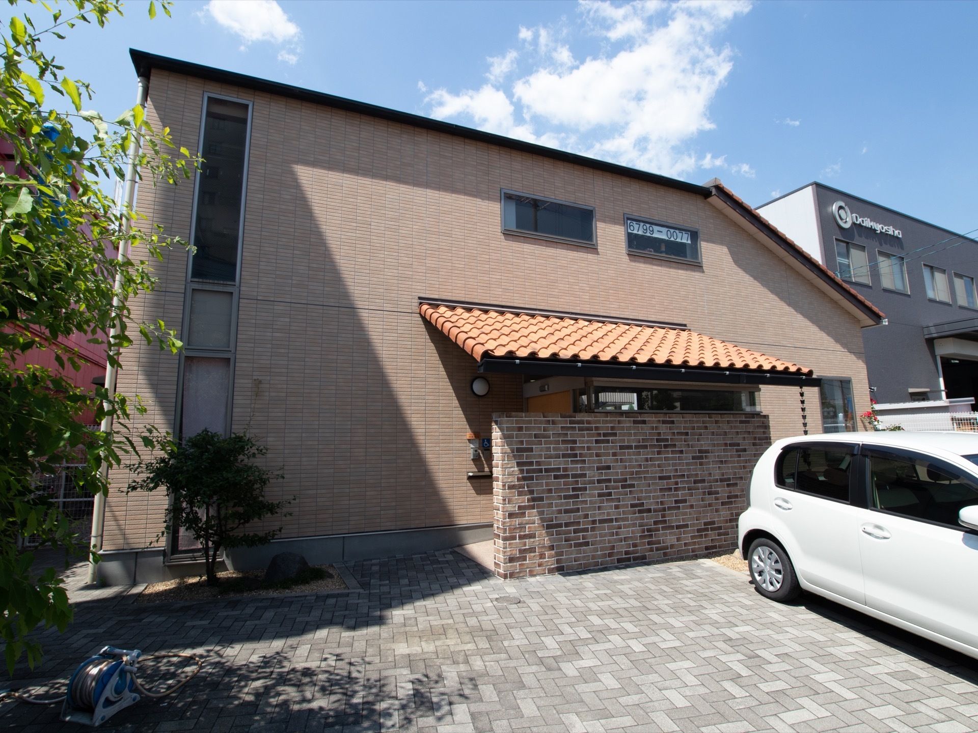 ２階建てでブラウンの建物があり、手前は駐車スペースになっている。周りには木々の植え込みが造られている。