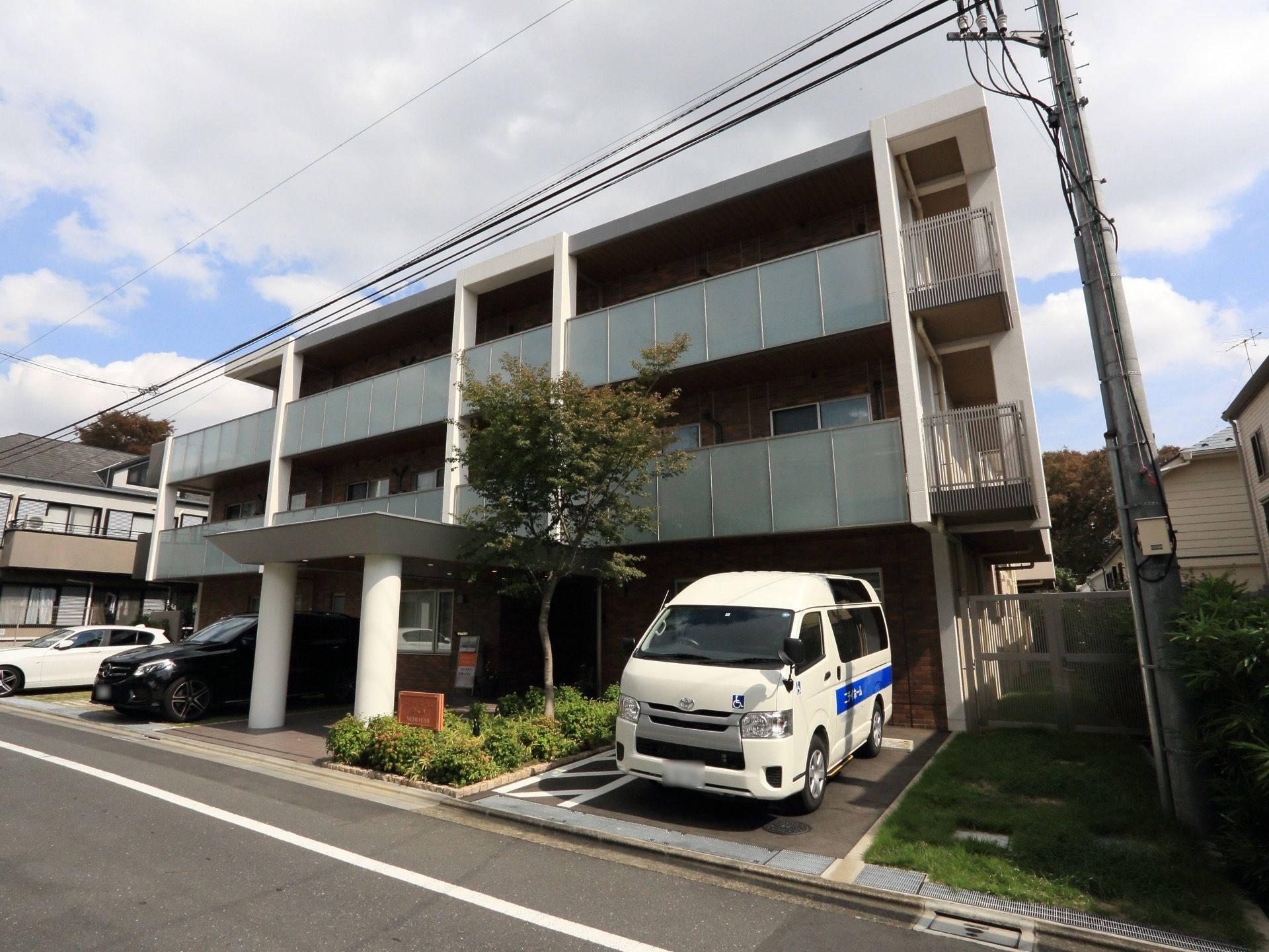 鉄筋コンクリート造りの地上３階建ての施設。建物の前には、自動車が３台停まっており、駐車スペースの脇に植栽が施されている。