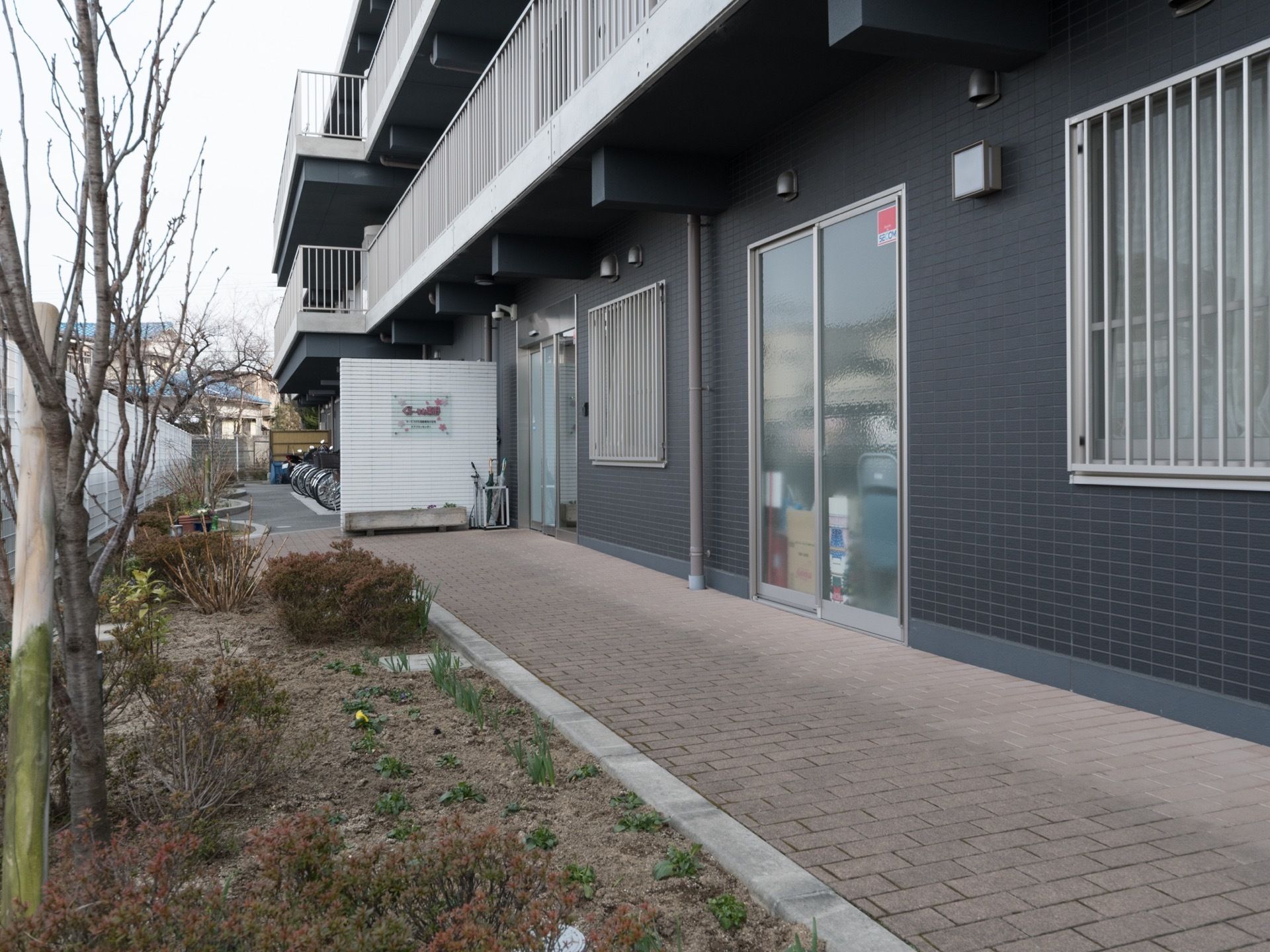エントランスの右側には、建物に沿ってタイルが敷かれた通路がある。フェンス際に花壇が設けられている。傘立てが玄関ドア横に置かれている。